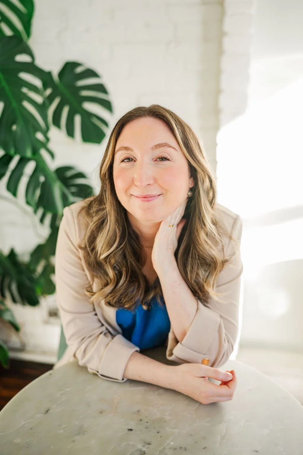 A woman with long wavy hair, smiling, resting her chin on her hand, sitting at a marble table with a large green leaf plant in the background, and bright natural light.