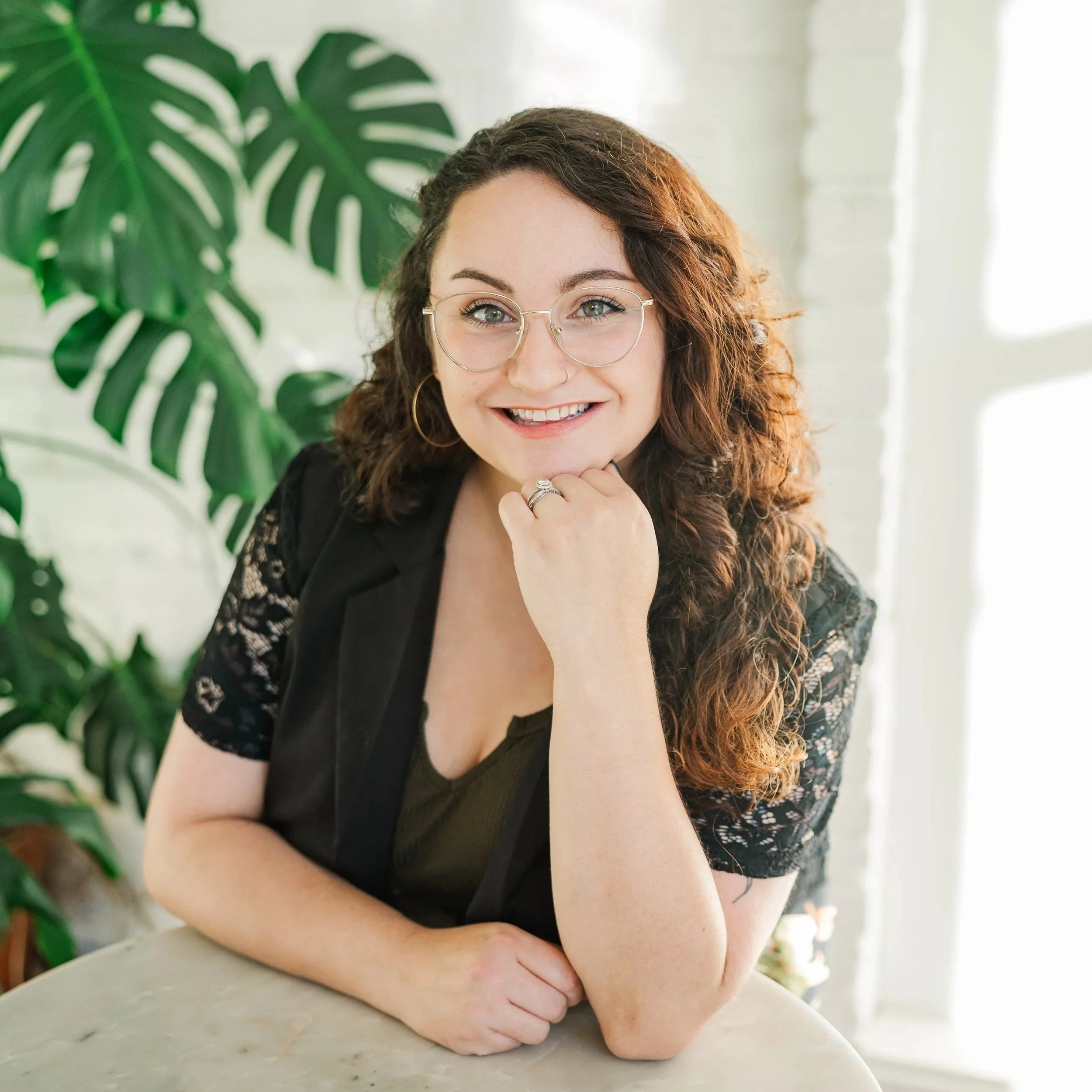 A woman with curly brown hair, glasses, and hoop earrings smiling at the camera, resting her chin on her hand, sitting at a marble table with a large green plant in the background.