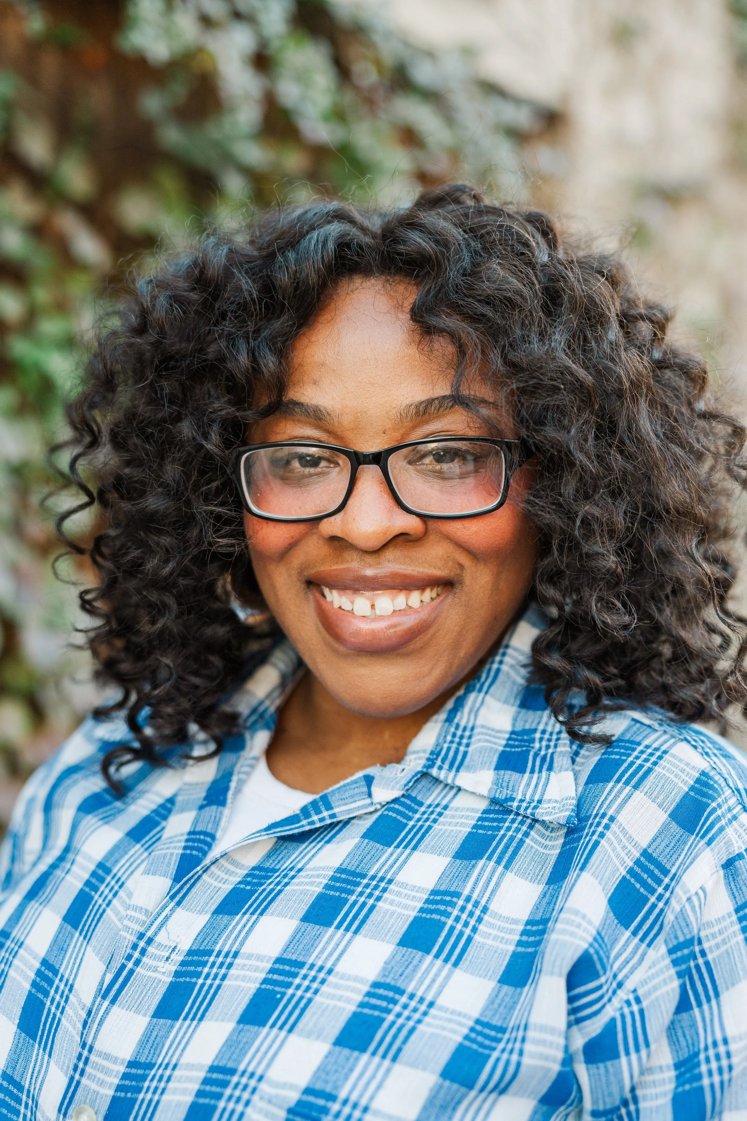Smiling woman with curly hair, glasses, and a plaid shirt outdoors.