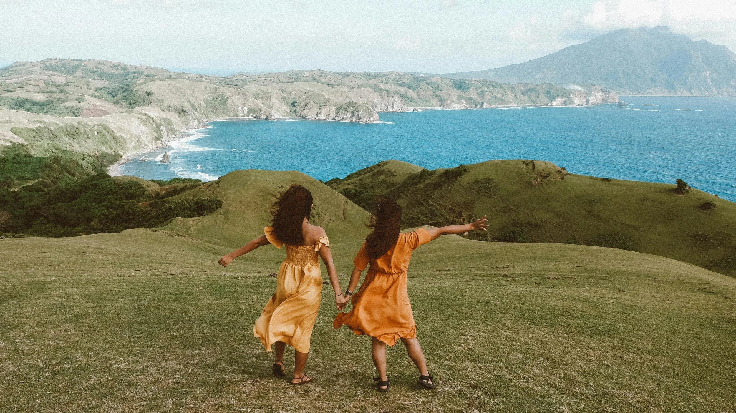 Two women wearing orange dresses holding hands and looking out to an ocean behind hills