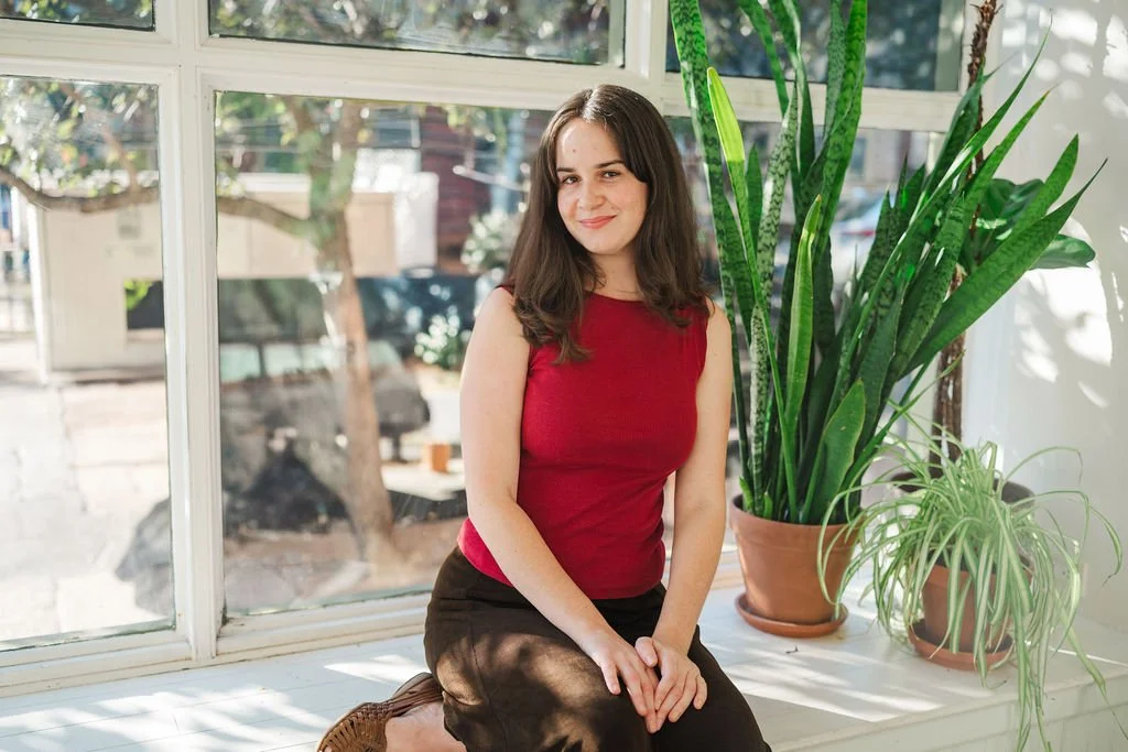 Young woman in a red sleeveless top and dark pants sitting by large indoor plants in front of a window.