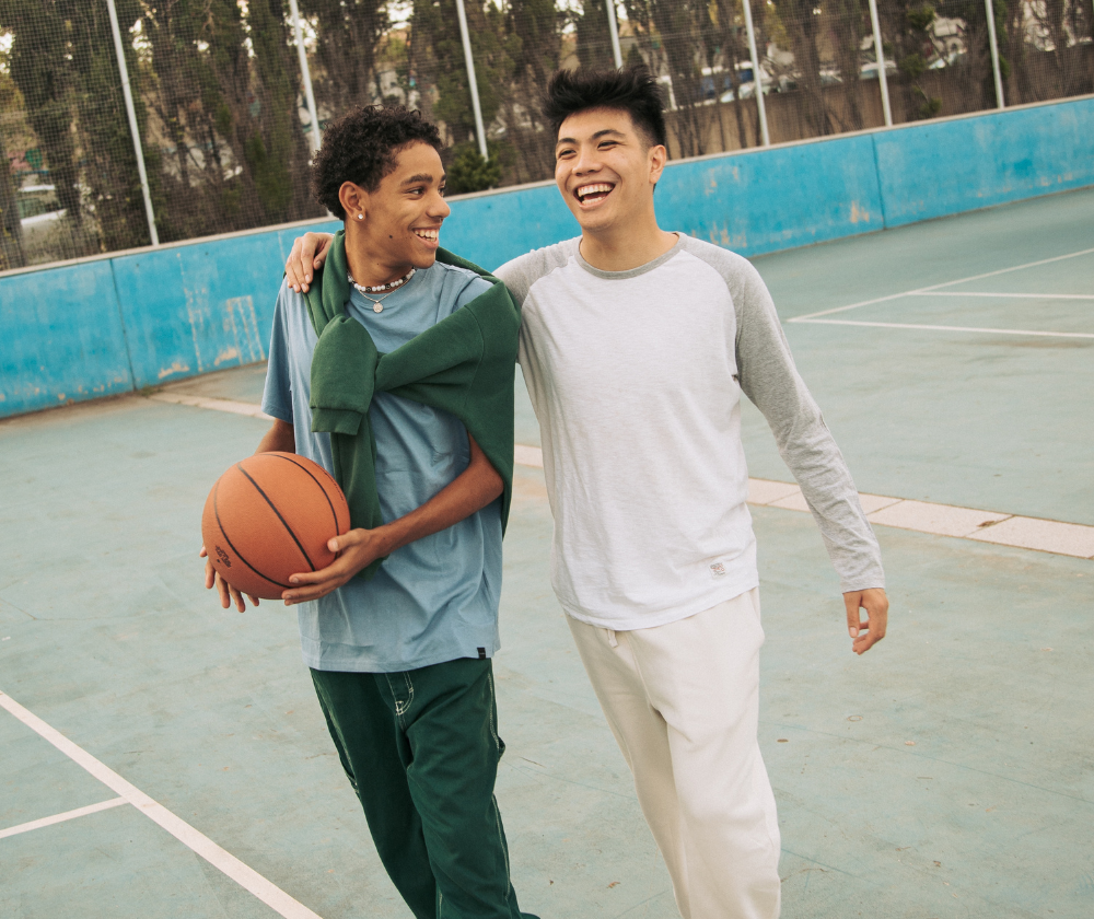 Two young men walking arm in arm on an outdoor basketball court, smiling and laughing, with one holding a basketball.