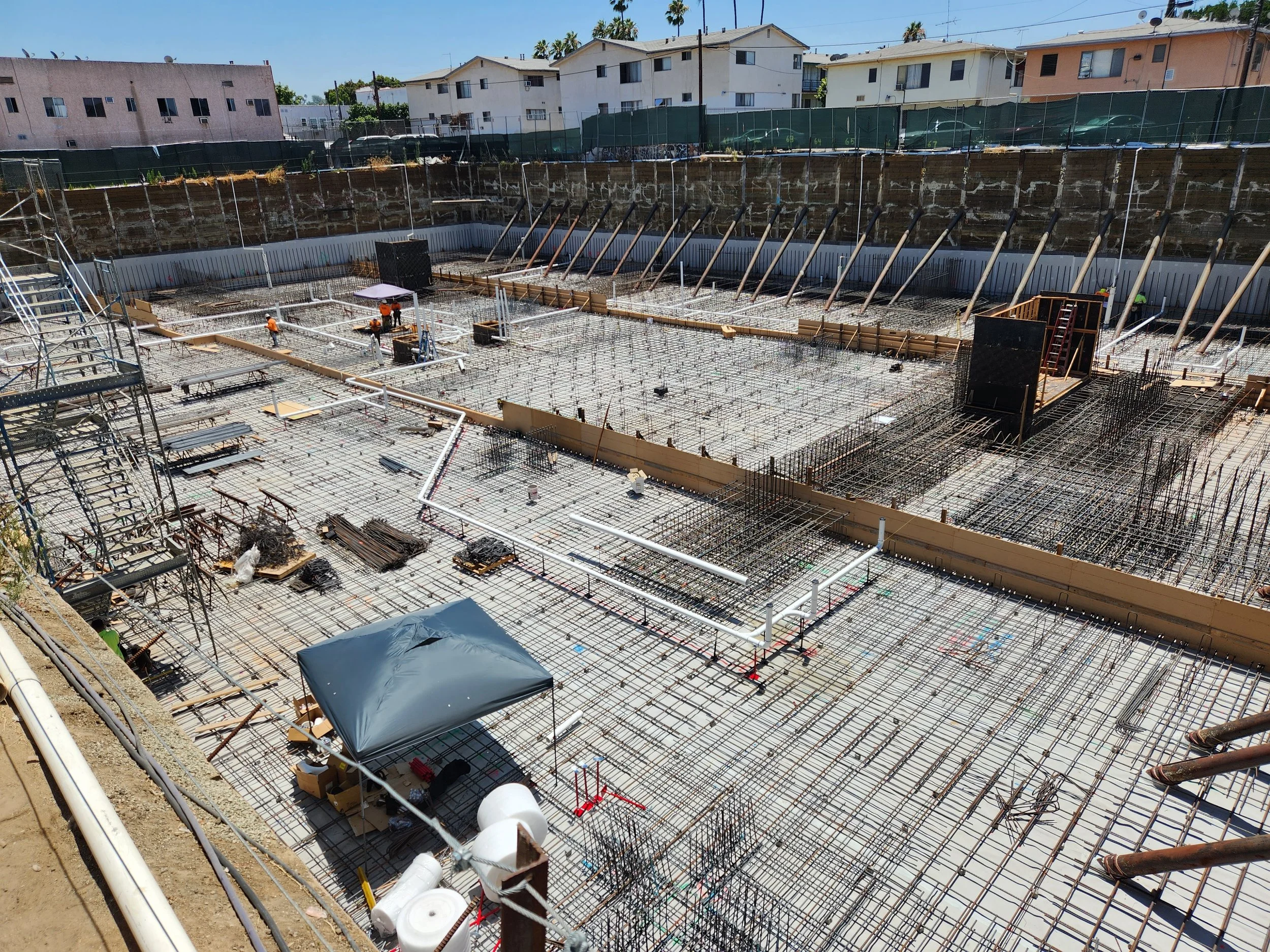 Construction site with workers and rebar for building a foundation, surrounded by residential houses and blue sky.