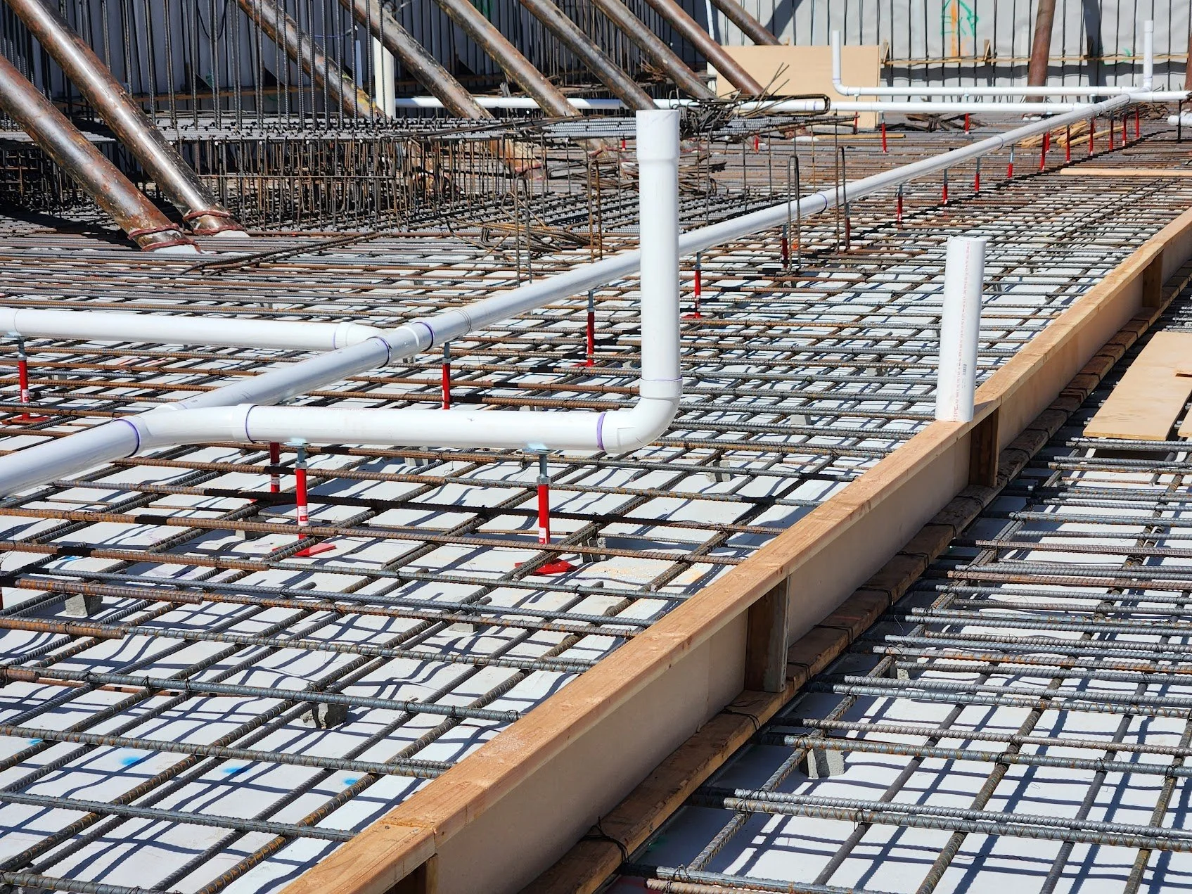Construction site with steel rebar grid and white plumbing pipes laid out on a wooden form for concrete pouring.