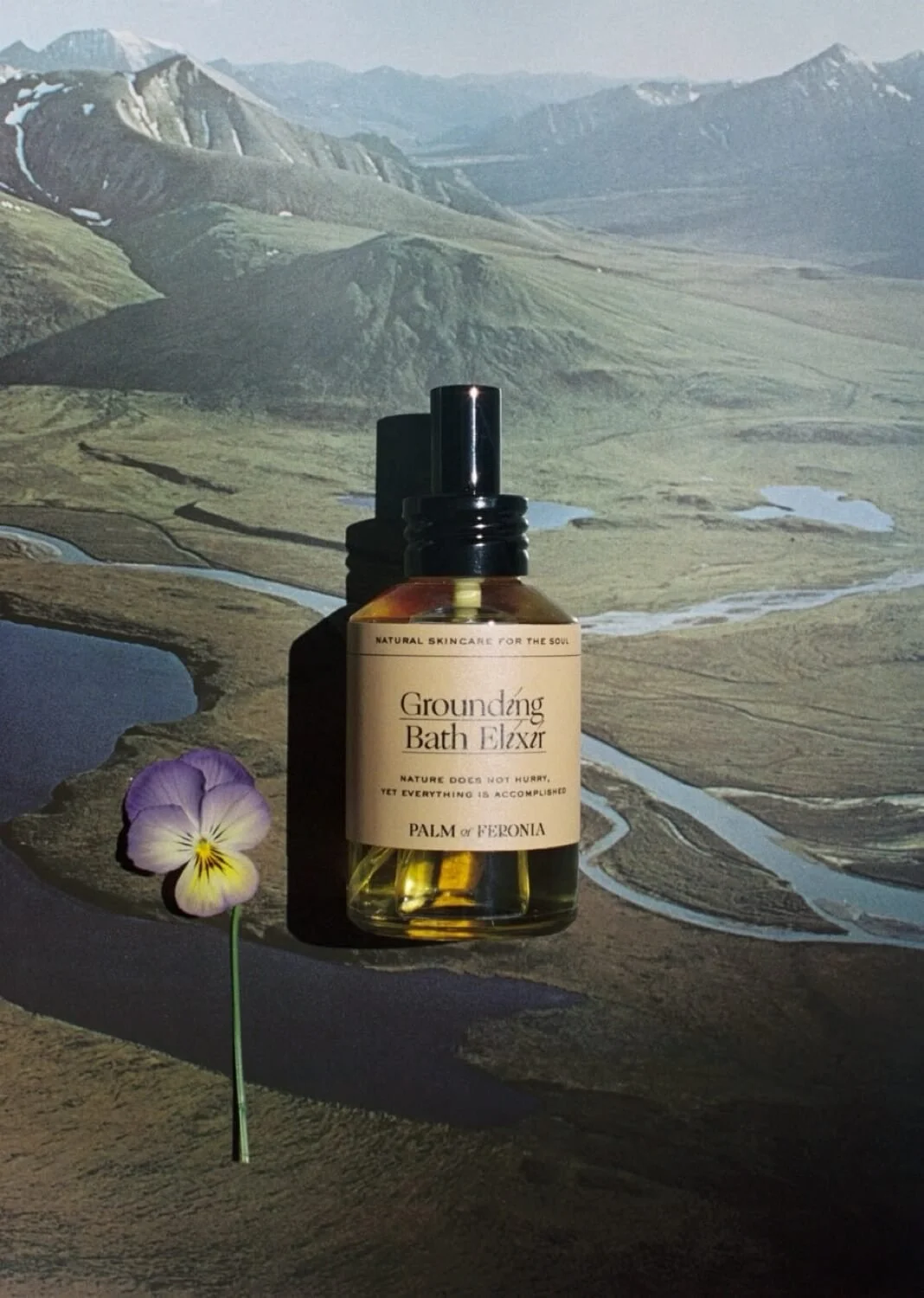 Grounding Bath Elixir by Palm of Feronia beside a pansy, set against a landscape background.