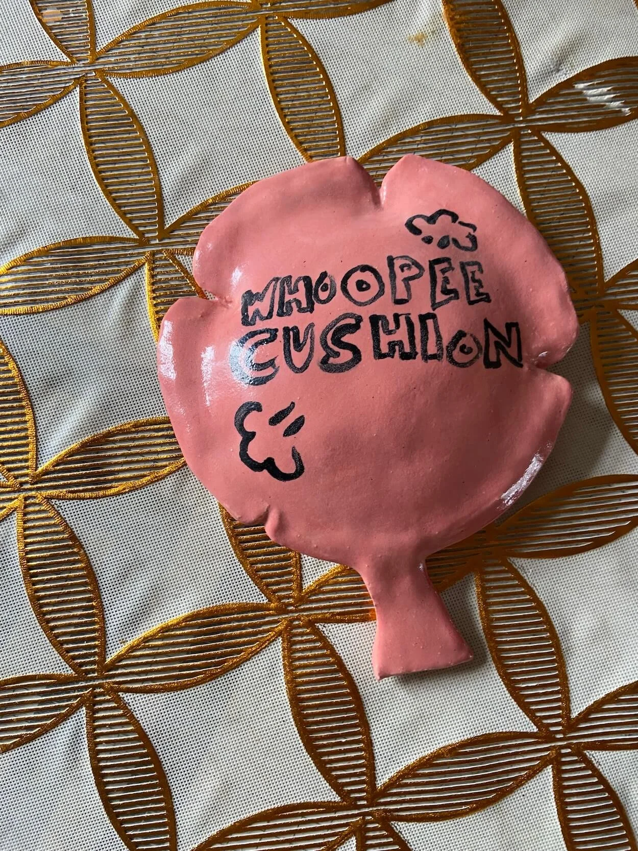 Ceramic whoopee cushion sculpture by Jonny Hughes displayed on a patterned plastic tablecloth in Alexandra Mann's home.