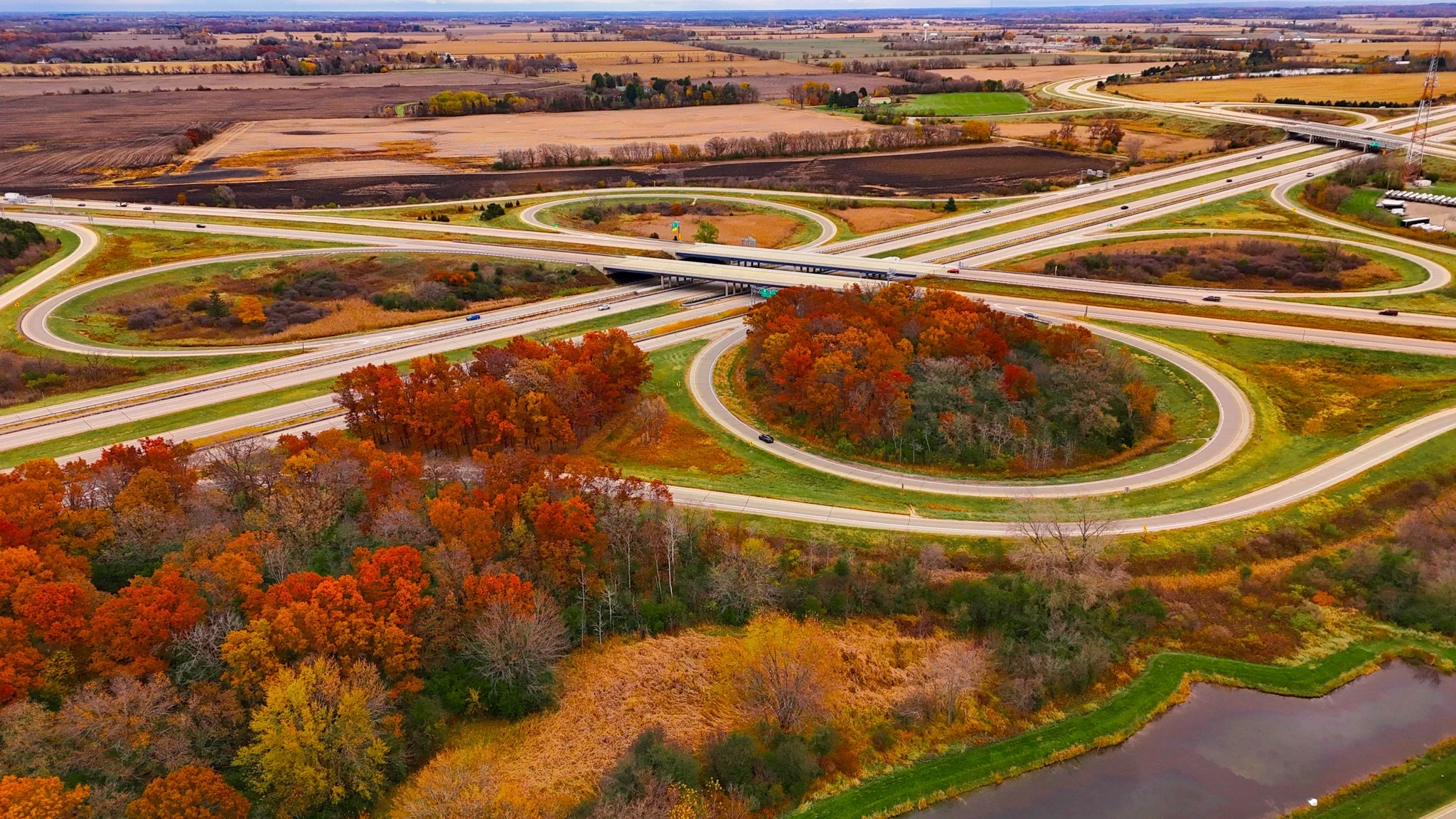An aerial view of a highway interchange with multiple looping roads and overpasses surrounded by colorful autumn trees, fields, and a small pond.