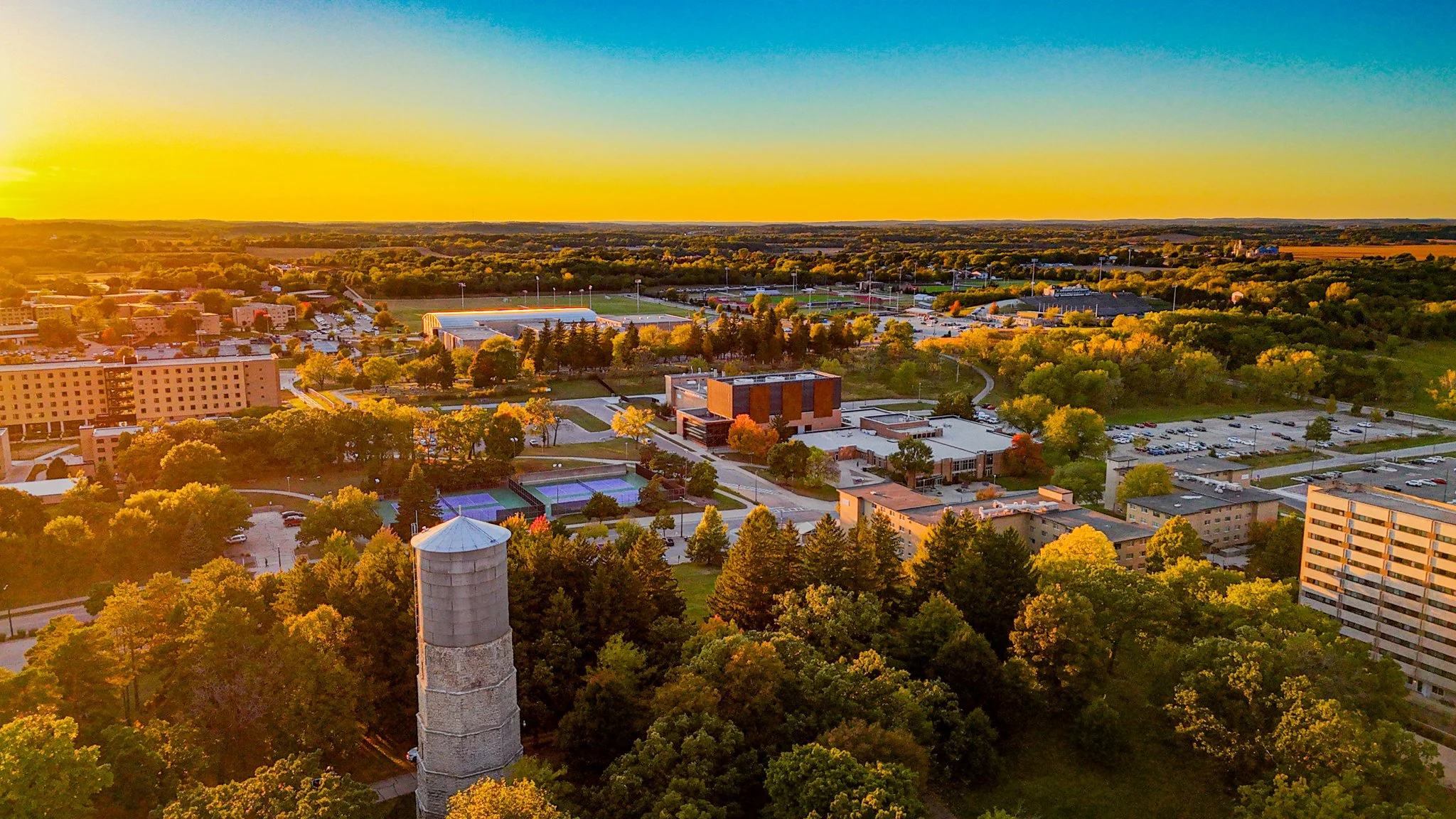 An aerial view of a college campus during sunset with trees, buildings, sports fields, and parking lots.