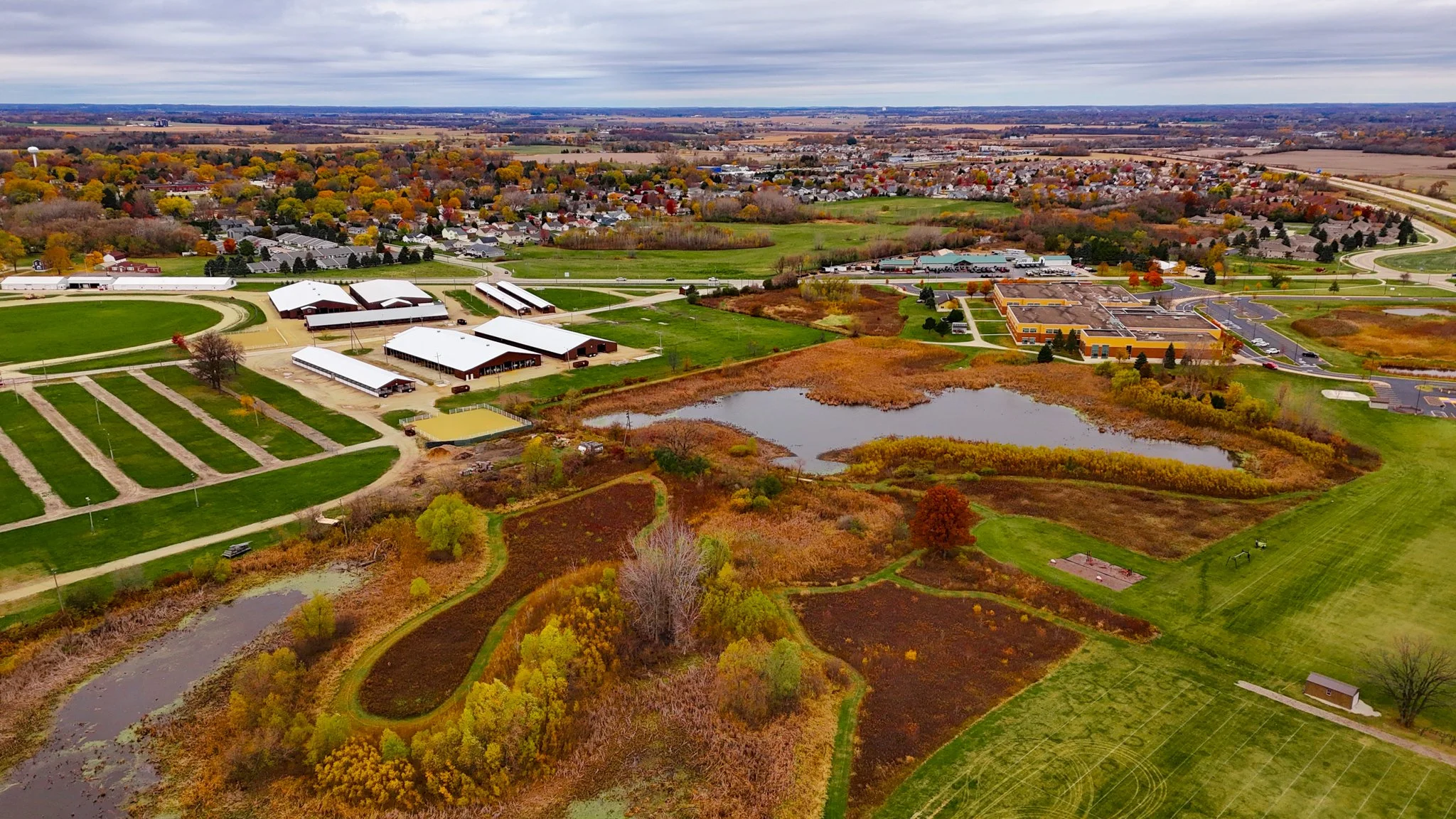 Aerial view of a community with a library, pond, farmland, and residential neighborhood surrounded by trees with fall foliage.
