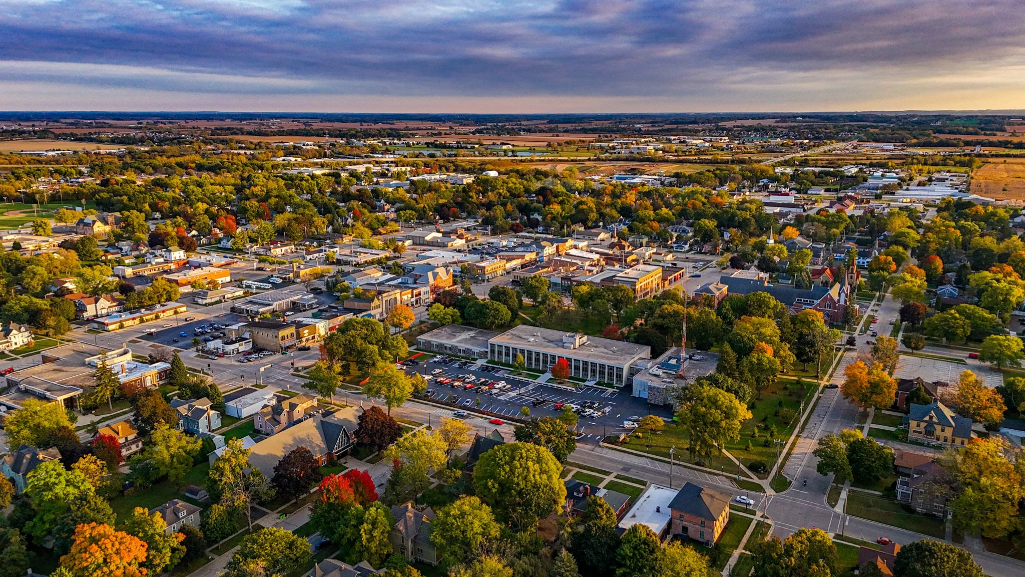 Aerial view of a small town with a downtown area, surrounded by trees with fall foliage, and open farmland in the background, under a partly cloudy sky at sunset.
