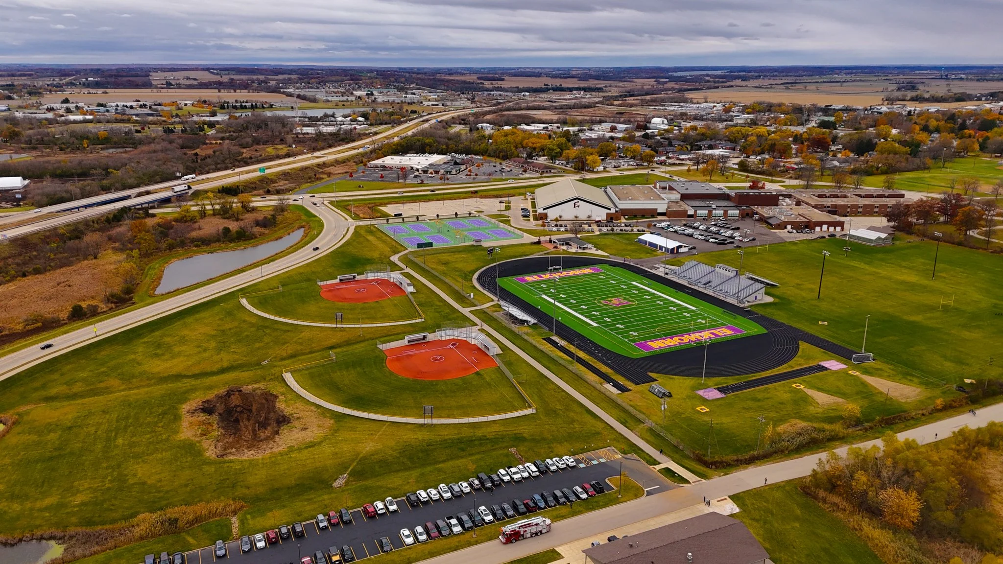 Aerial view of a school campus with a football field, multiple baseball and tennis courts, parking lots, and surrounding roads and nature.
