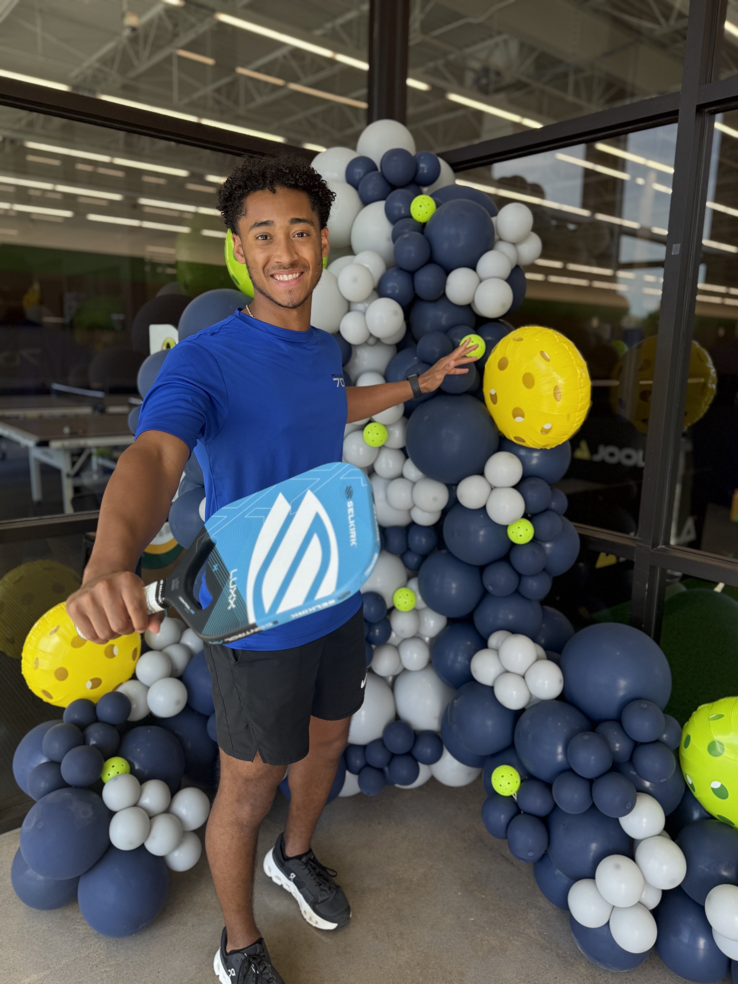 A young man in a blue t-shirt and black shorts holding a pickleball paddle, standing in front of a balloon display with assorted white, blue, and yellow balloons, some shaped like pickleballs and tennis balls, inside a sporting goods store.