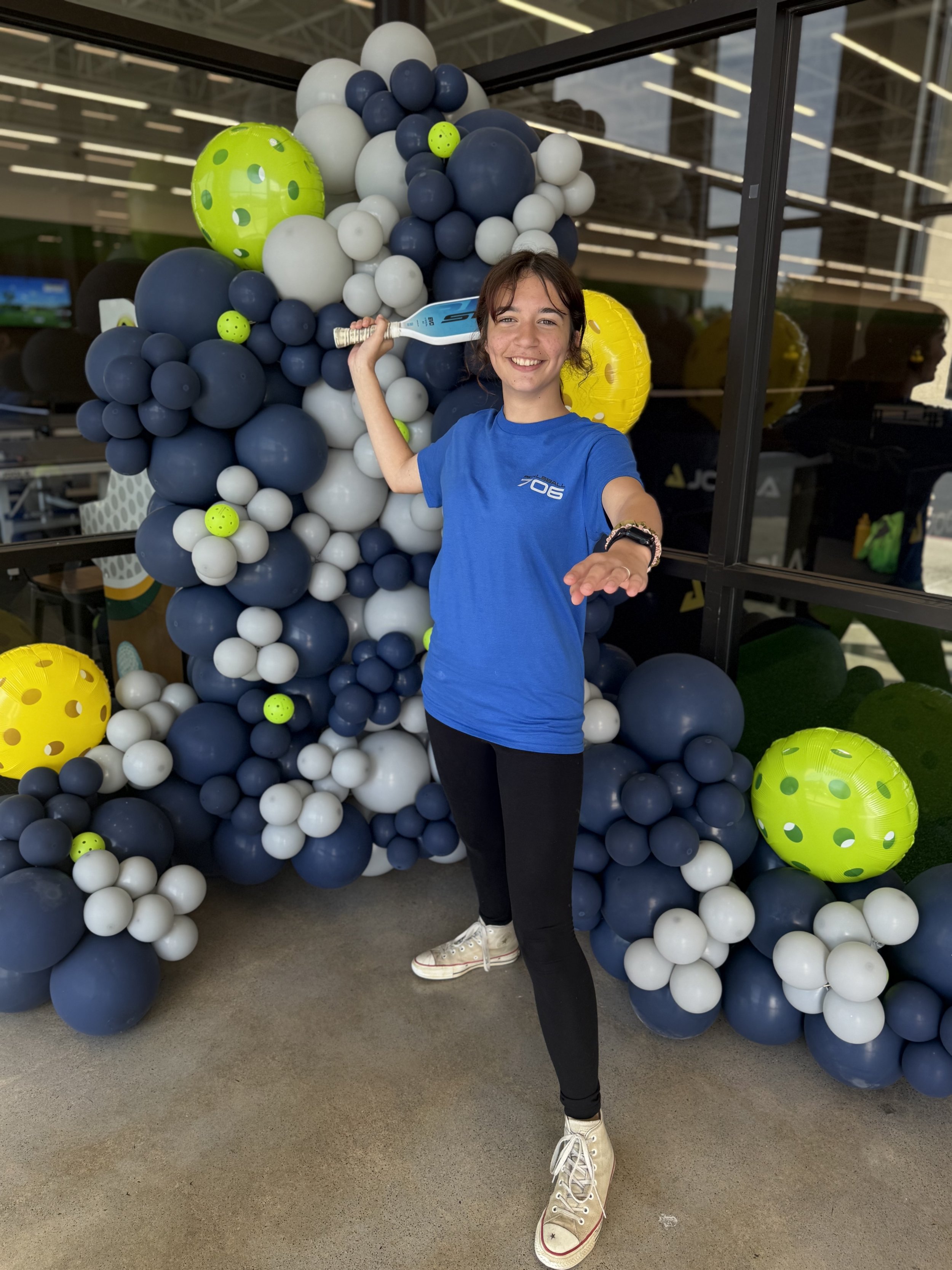 A young woman posing with an arm extended, holding a toy tennis racket, standing in front of a large balloon decoration featuring blue, white, yellow, and green balloons.