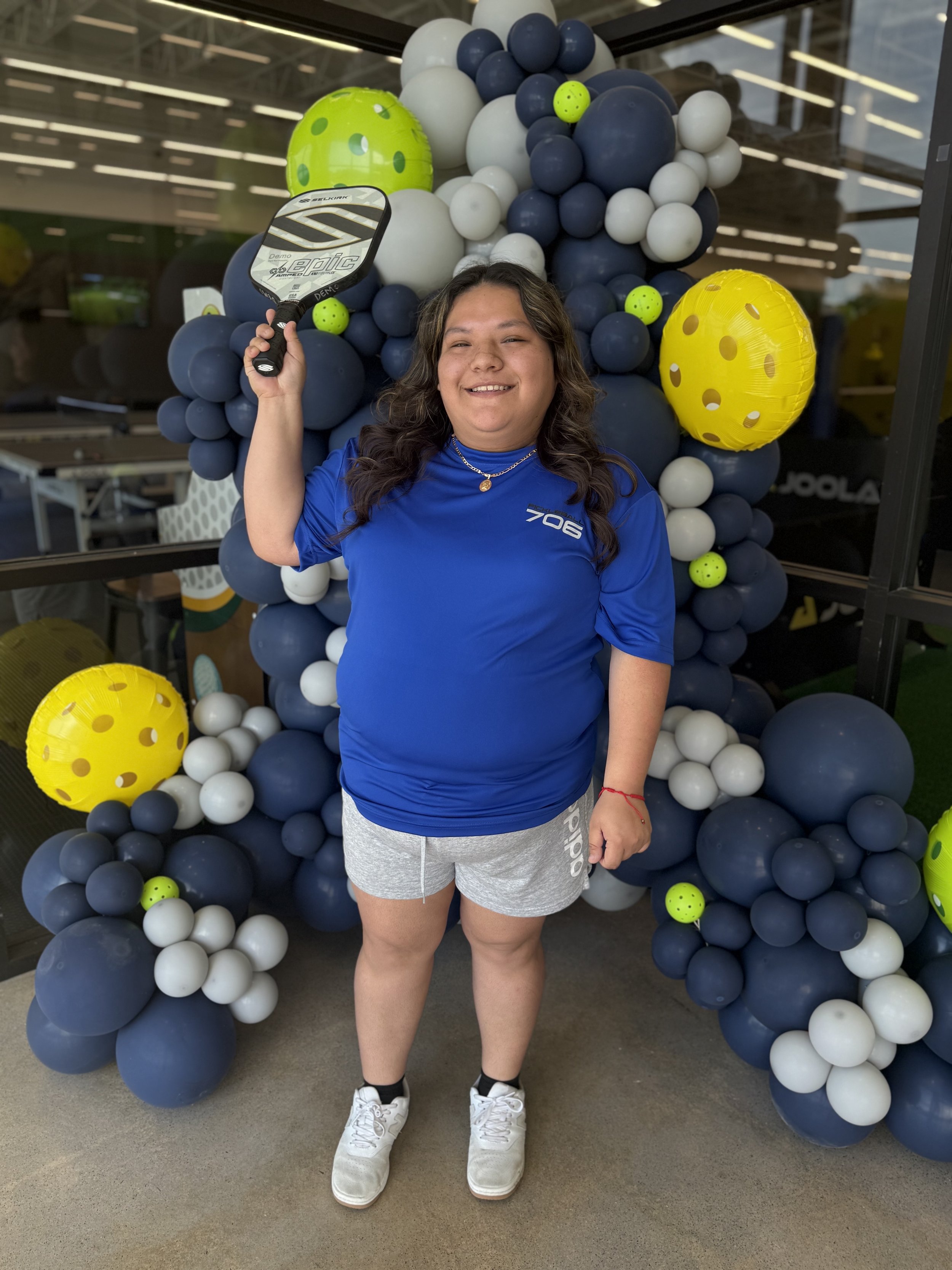 A young woman standing in front of a balloon display, holding a pickleball paddle, smiling at the camera, wearing a blue t-shirt and gray shorts.