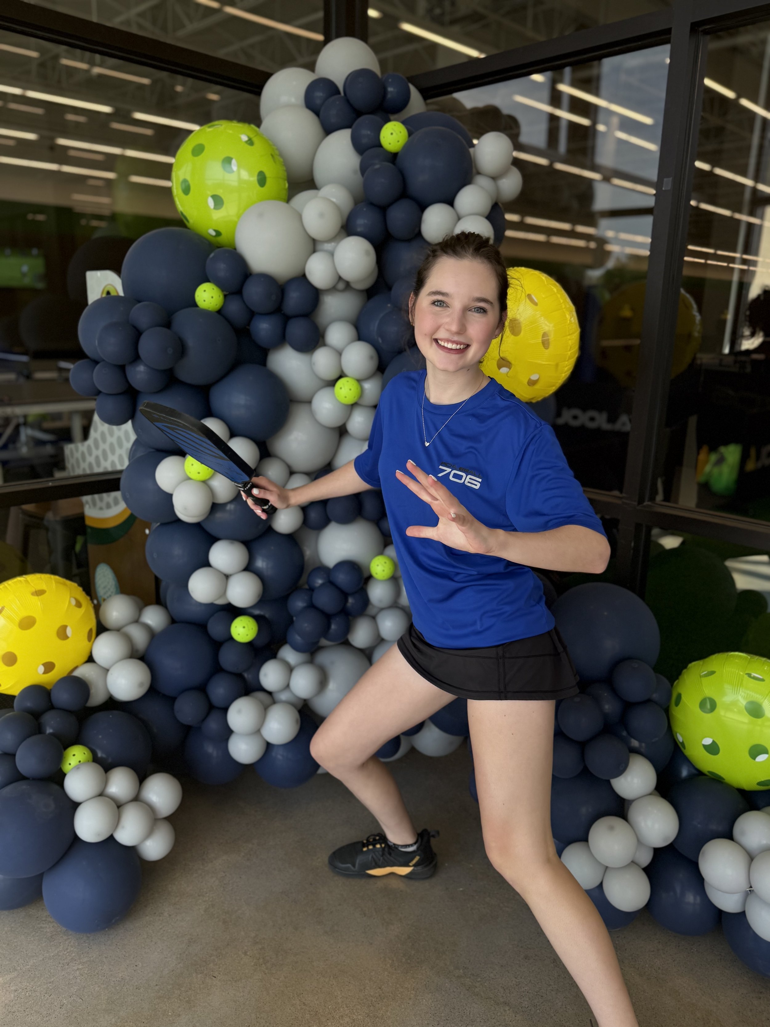 Young woman in a blue sports shirt and black shorts posing playfully in front of a large balloon arrangement with navy blue, white, and yellow balloons, some of which are designed to look like tennis balls.