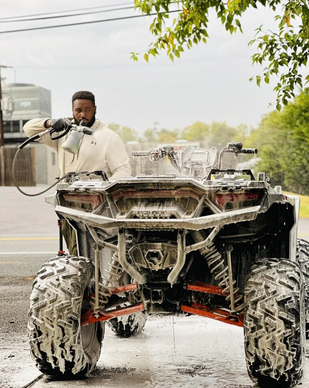 A man washing a damaged, dusty Polaris off-road vehicle with a pressure washer on a street.