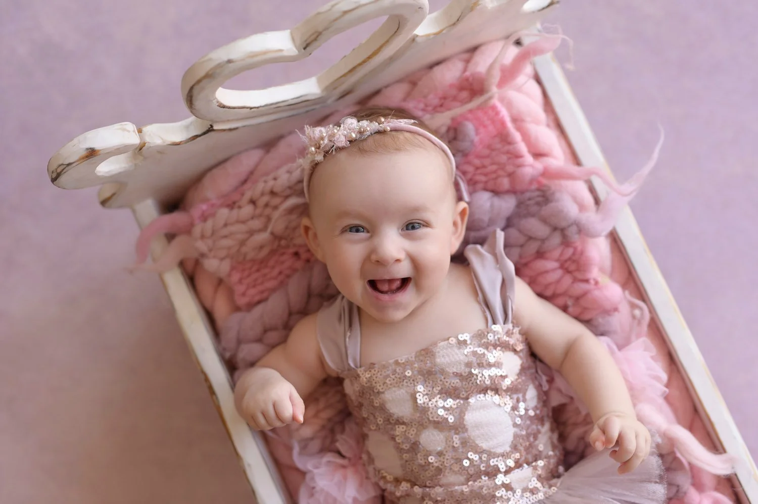 Smiling baby in a pink sequin dress and headband lying in a decorative wooden cradle with pink knitted blanket.