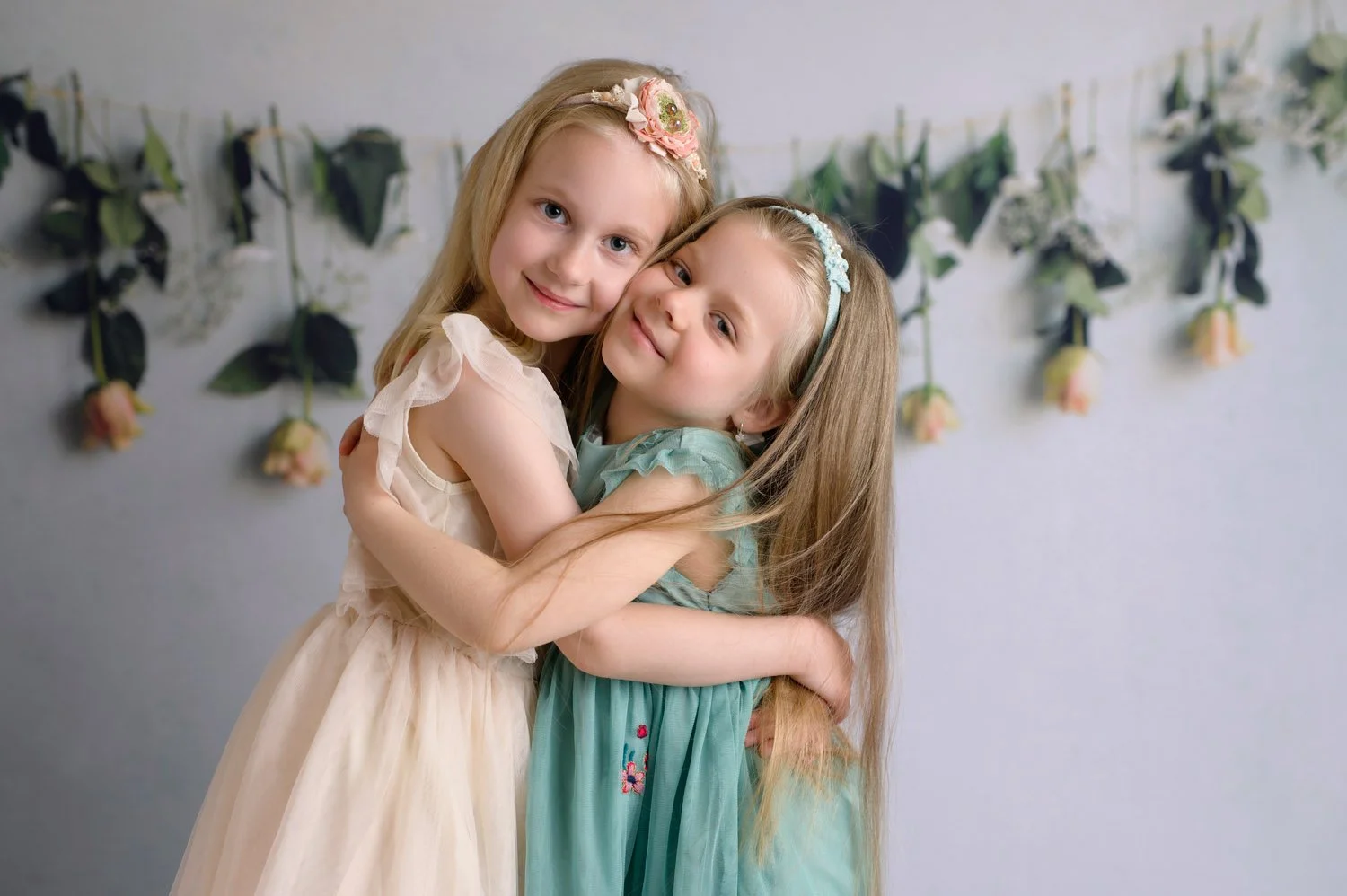 Two young girls hugging, wearing pastel dresses, with a floral garland backdrop. - kids photography studio in LJubljana