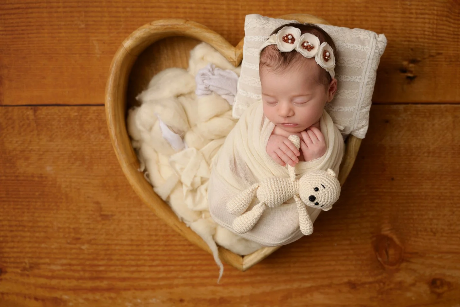Sleeping newborn wrapped in a cream blanket, lying in a heart-shaped wooden bowl with a knitted teddy bear, set on a wooden background.