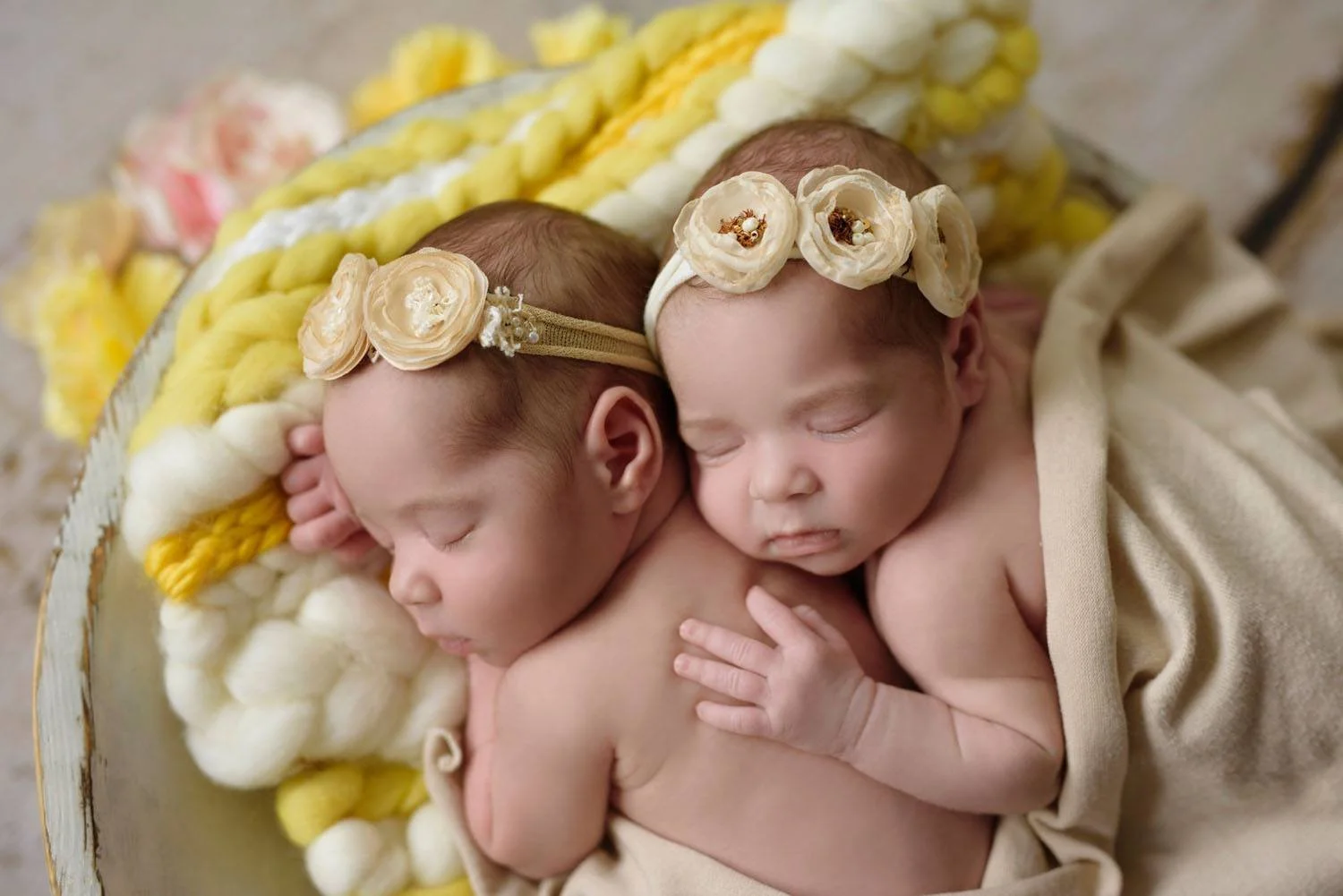 Newborn twins sleeping together, adorned with flower headbands, wrapped in a soft beige blanket with a yellow and white wool backdrop.