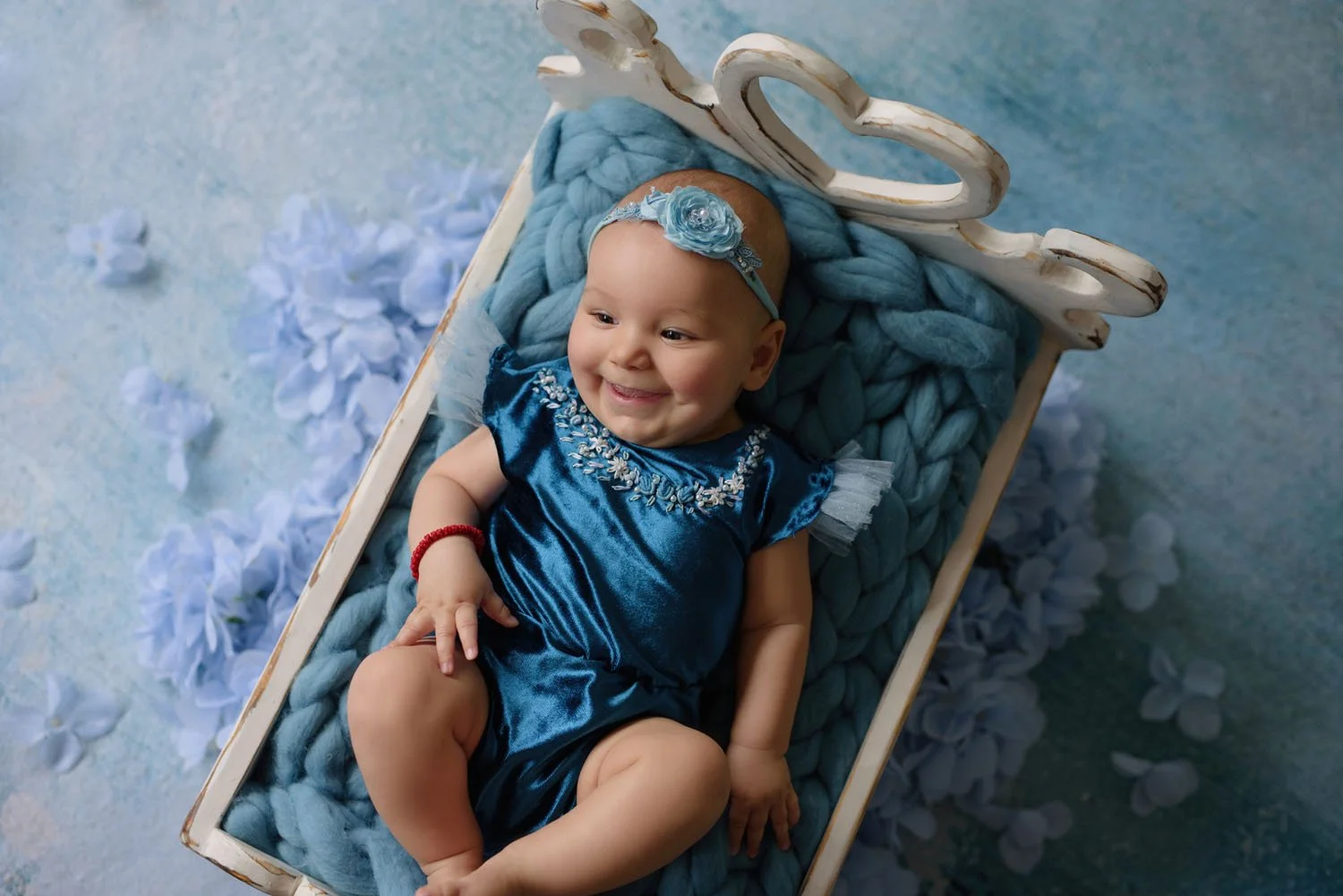 Smiling baby in blue dress lying on fluffy blanket in white cradle
