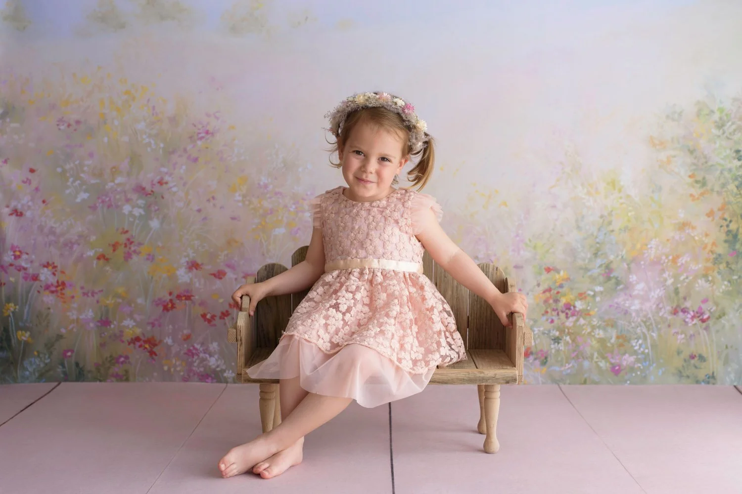Young girl in pink lace dress sitting on wooden bench with floral background.