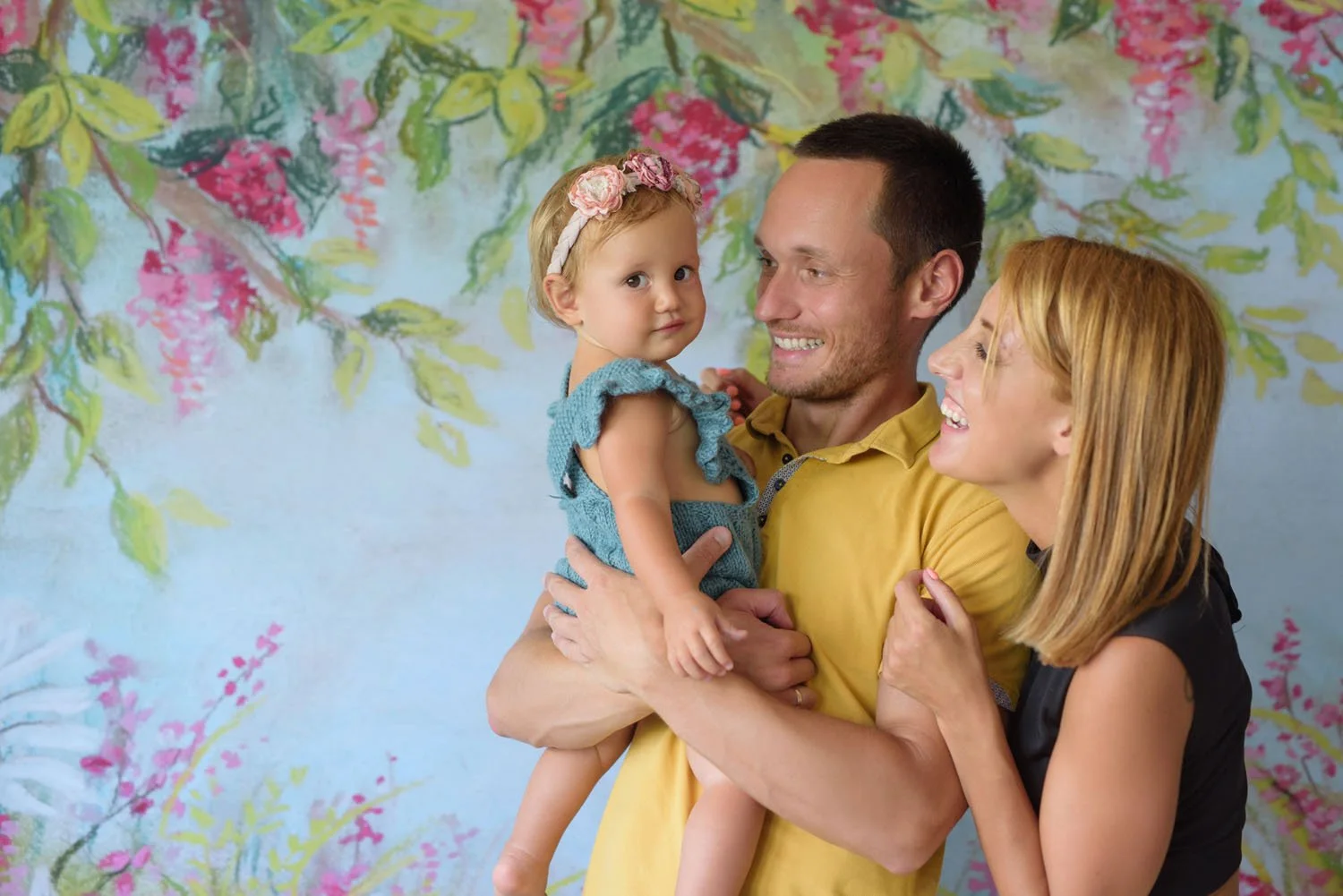 Družinska fotografija – nasmejan oče drži majhnega otroka v naročju, mama stoji ob njiju, v ozadju pa je cvetlično poslikano platno.