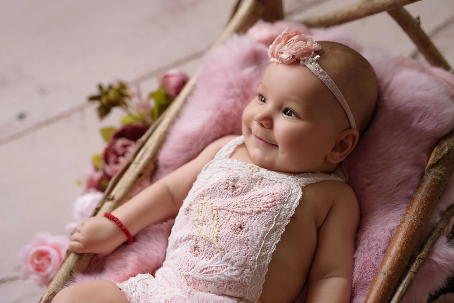 A smiling baby wearing a pink headband with a flower, lying on a fluffy pink blanket in a wooden basket, surrounded by pink and green floral decorations.