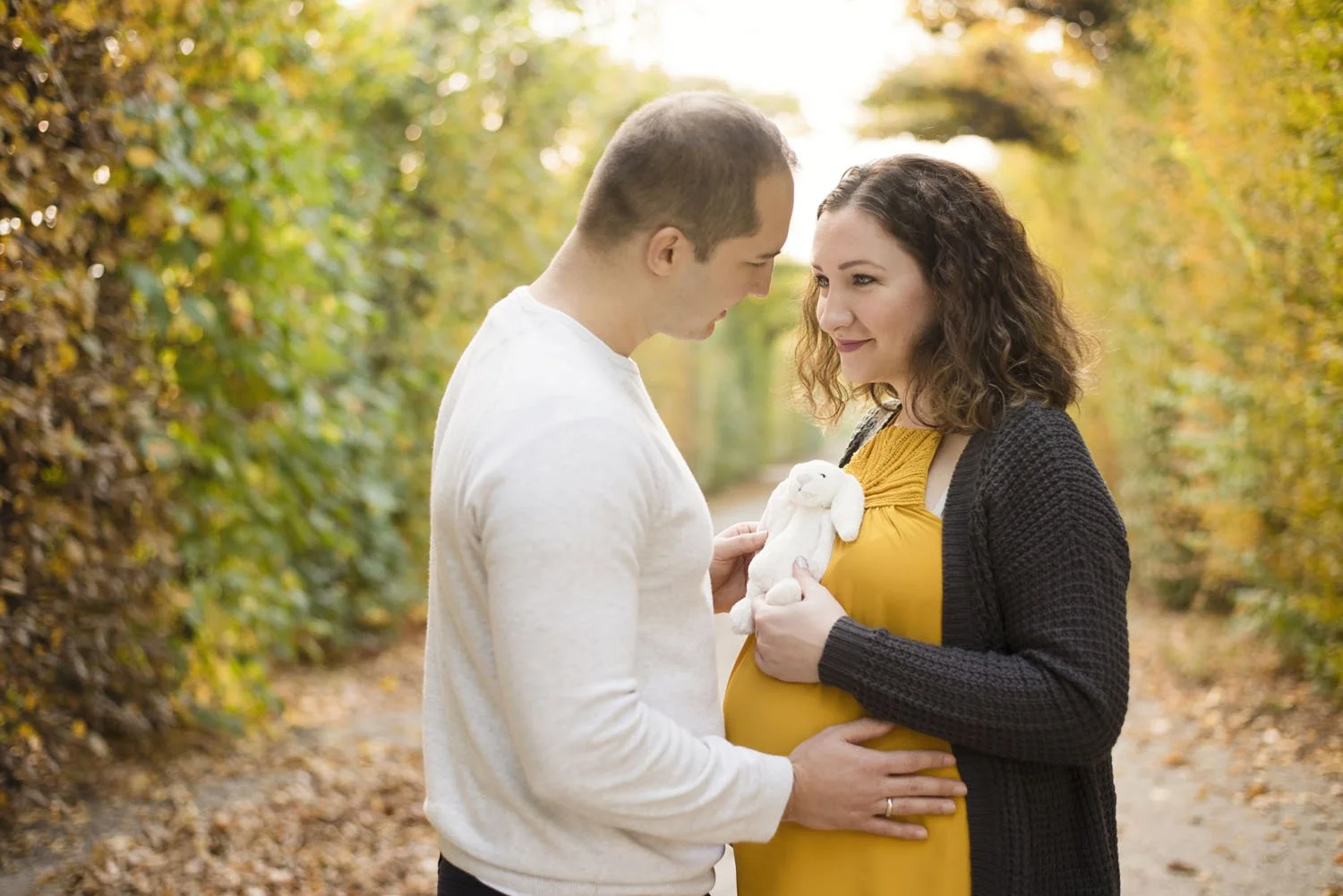 Pregnant couple embracing on a leafy path, woman holding a stuffed rabbit toy over her belly. Maternity photographer in Slovenia.