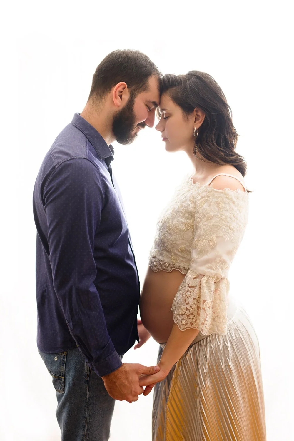Pregnant couple standing face to face, holding hands, with foreheads touching, in a bright, backlit setting.