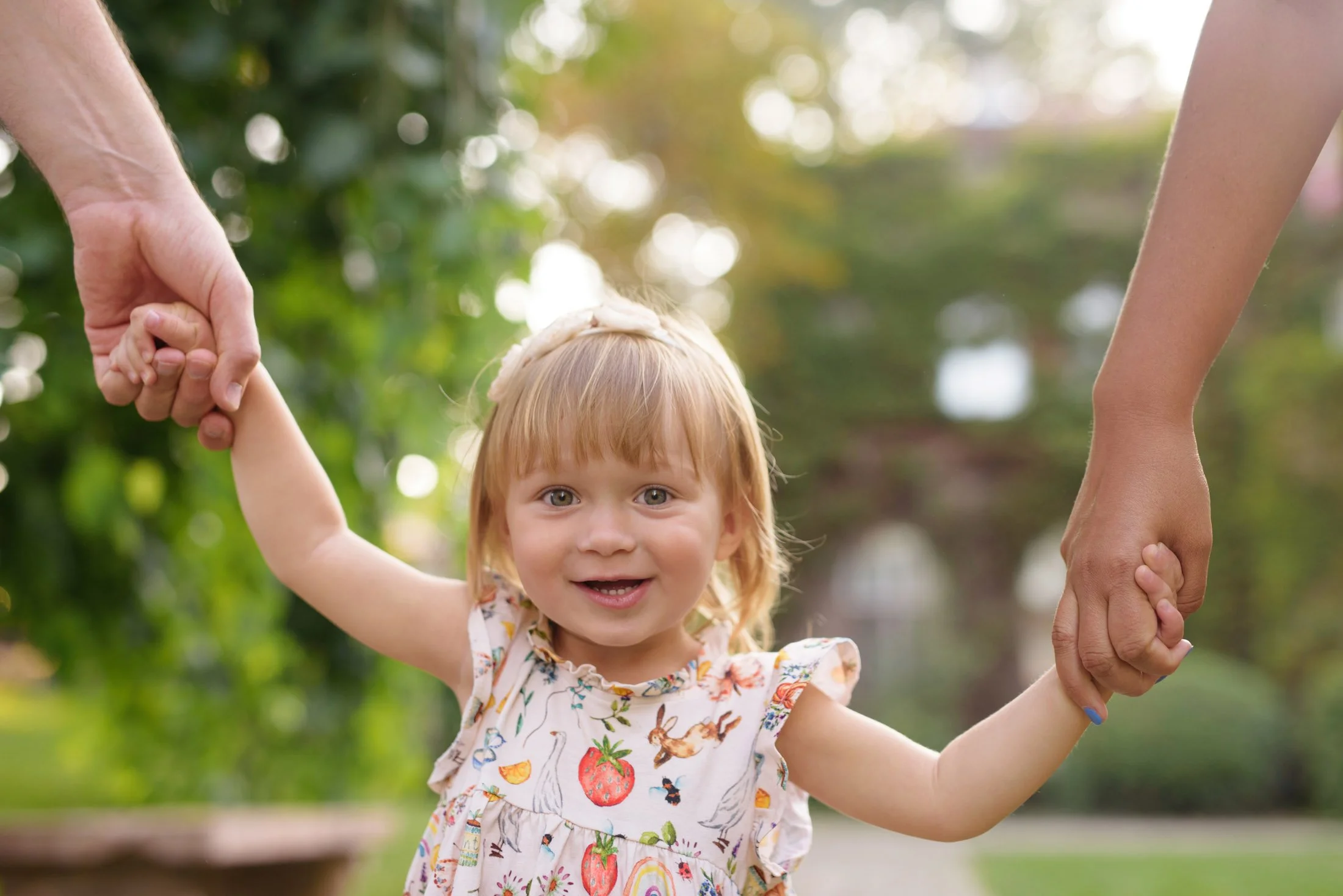 A smiling child holding hands with two adults, standing in a garden with blurred greenery in the background.
