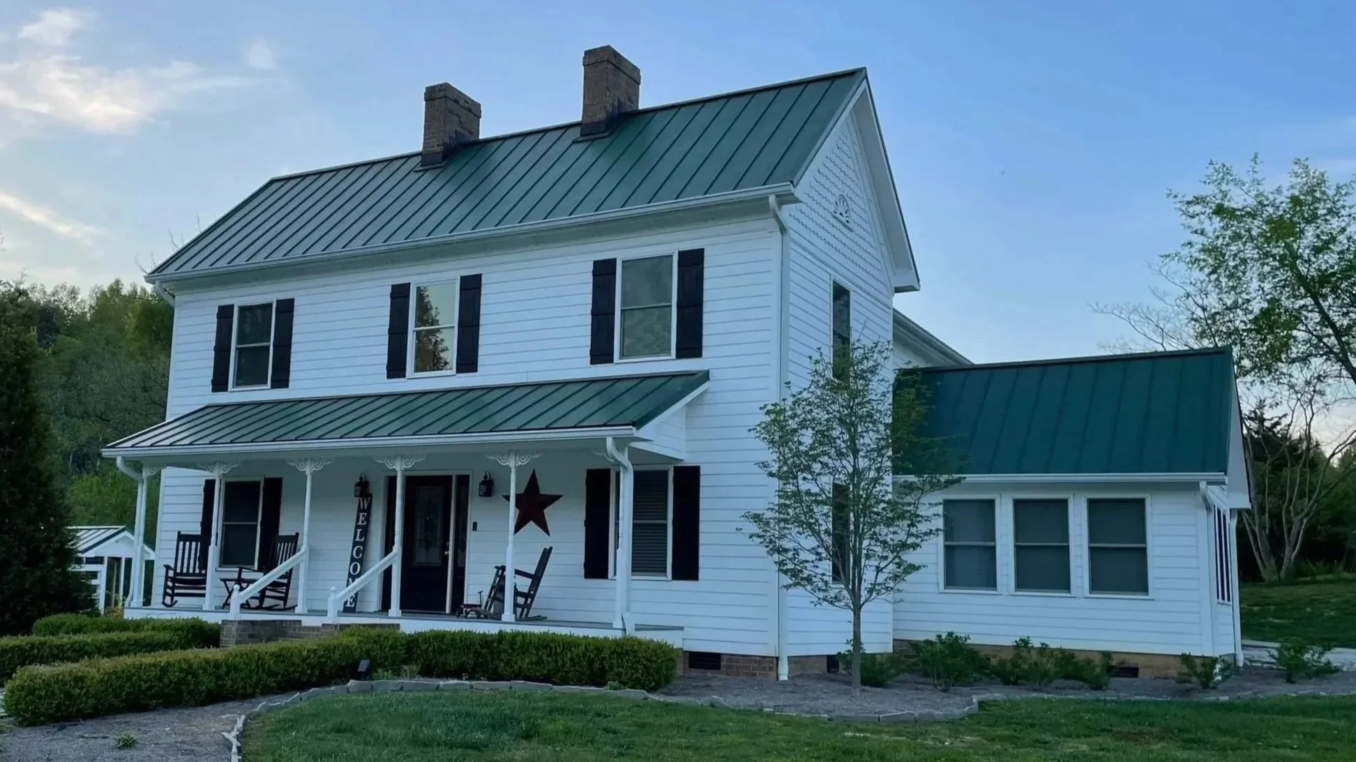 A white, two-story Smoky Mountain vacation rental with a steep green metal roof and black shutters on the windows. The house has a front porch with decorative supports, a swing, and a welcome sign.