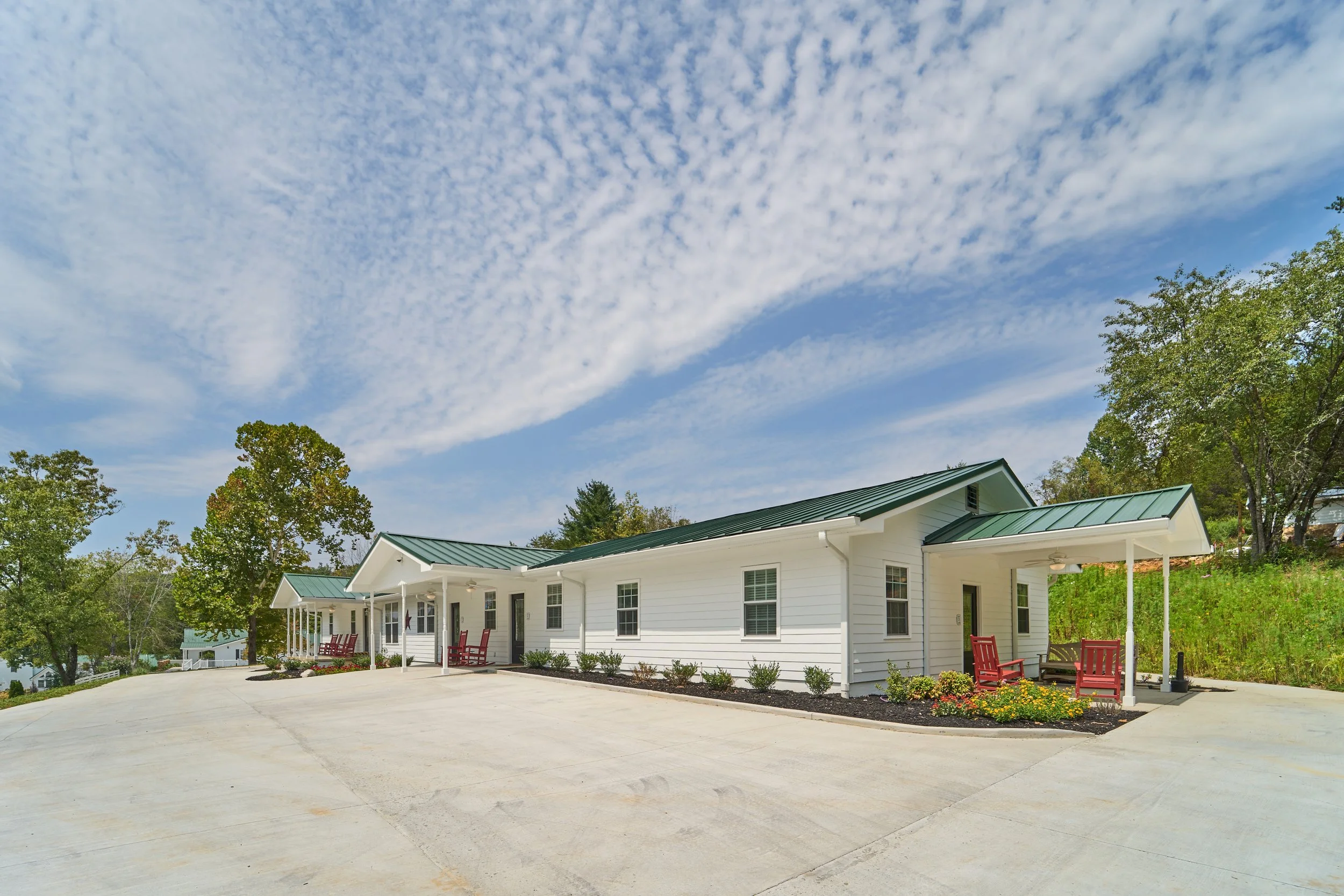 White modern building with a green metal roof, porch with red chairs, and landscaped lawn, under a blue sky with scattered clouds and surrounded by green trees.