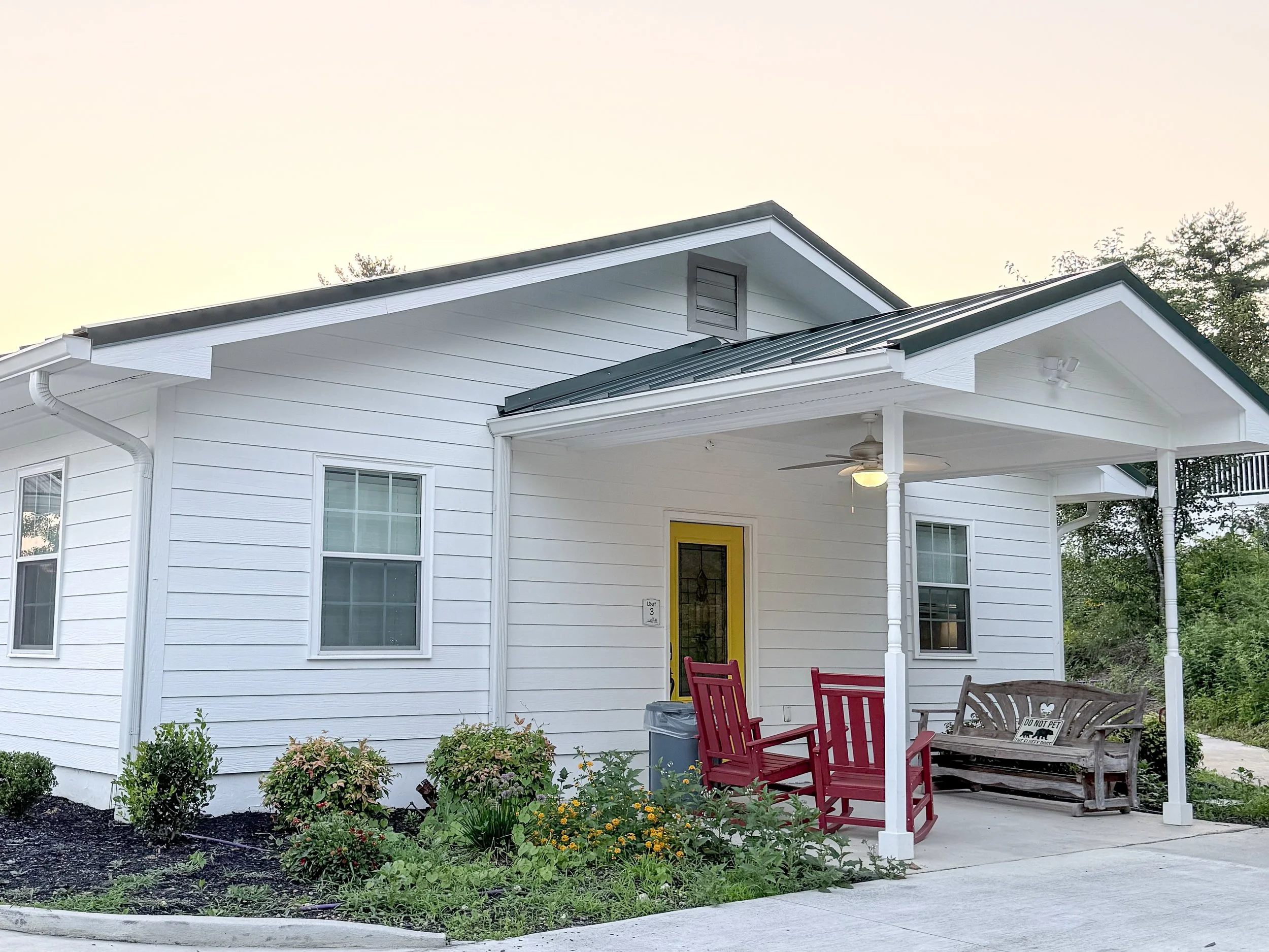Single-story white house with green roof and yellow door, featuring a covered porch with red rocking chairs and a wooden glider, surrounded by landscaped gardens, a wildflower bed, and bright skies at sunset.