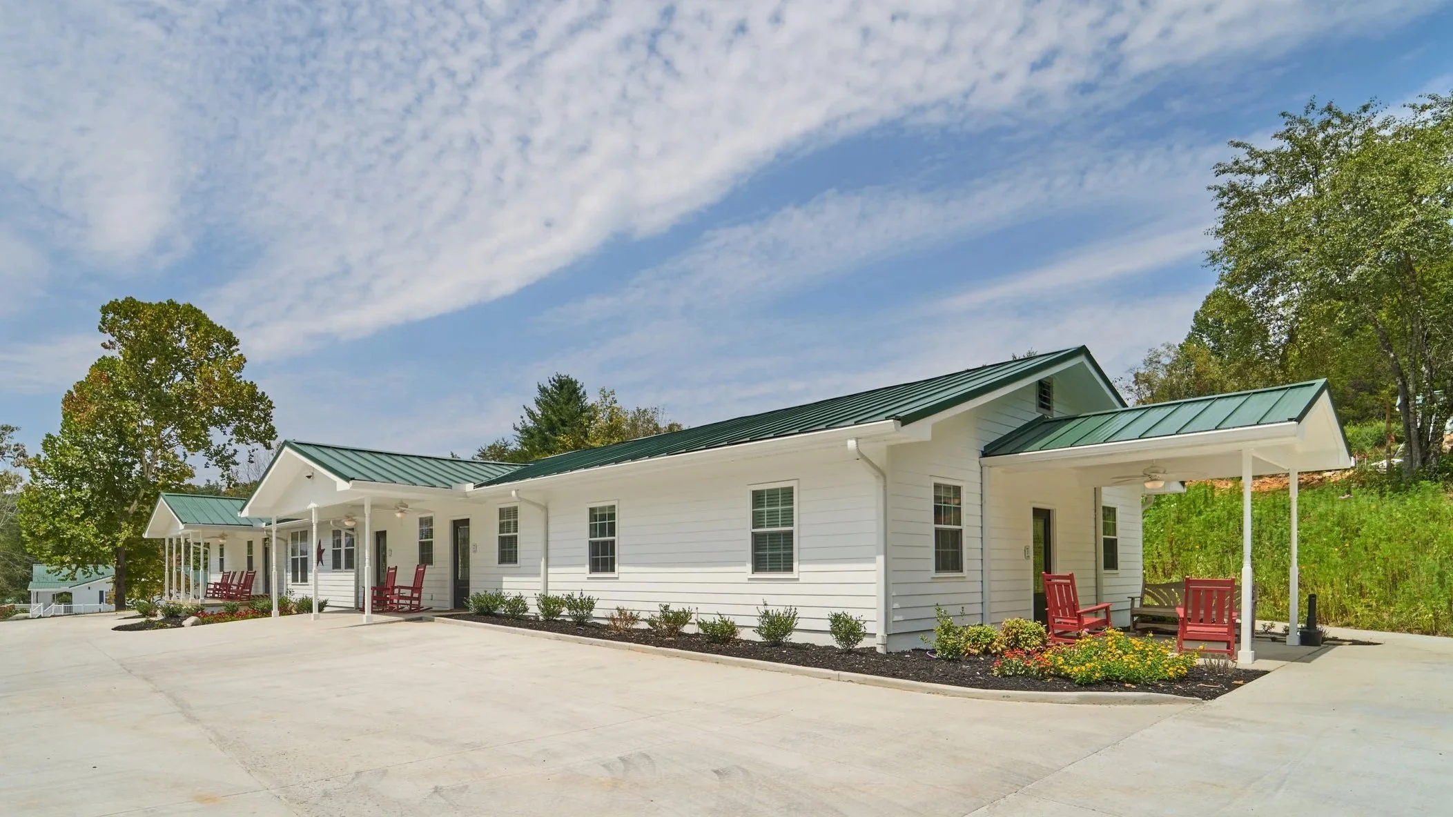 A multiunit property with white siding and green roof.  Ample flat parking is visible, as is the wildflower patch behind the property.