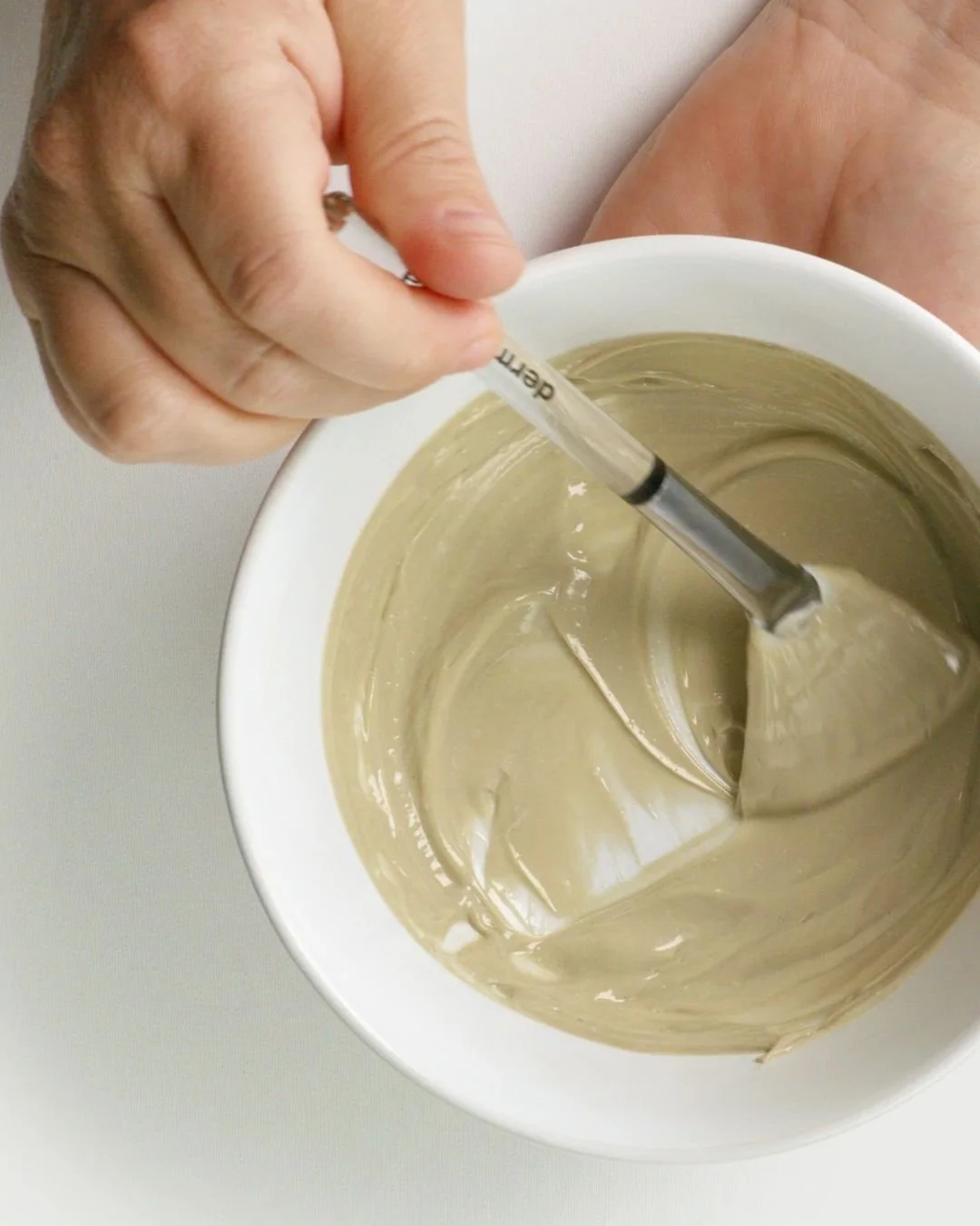 A hand stirring a light brown, smooth, creamy substance in a white bowl with a glass stir stick.