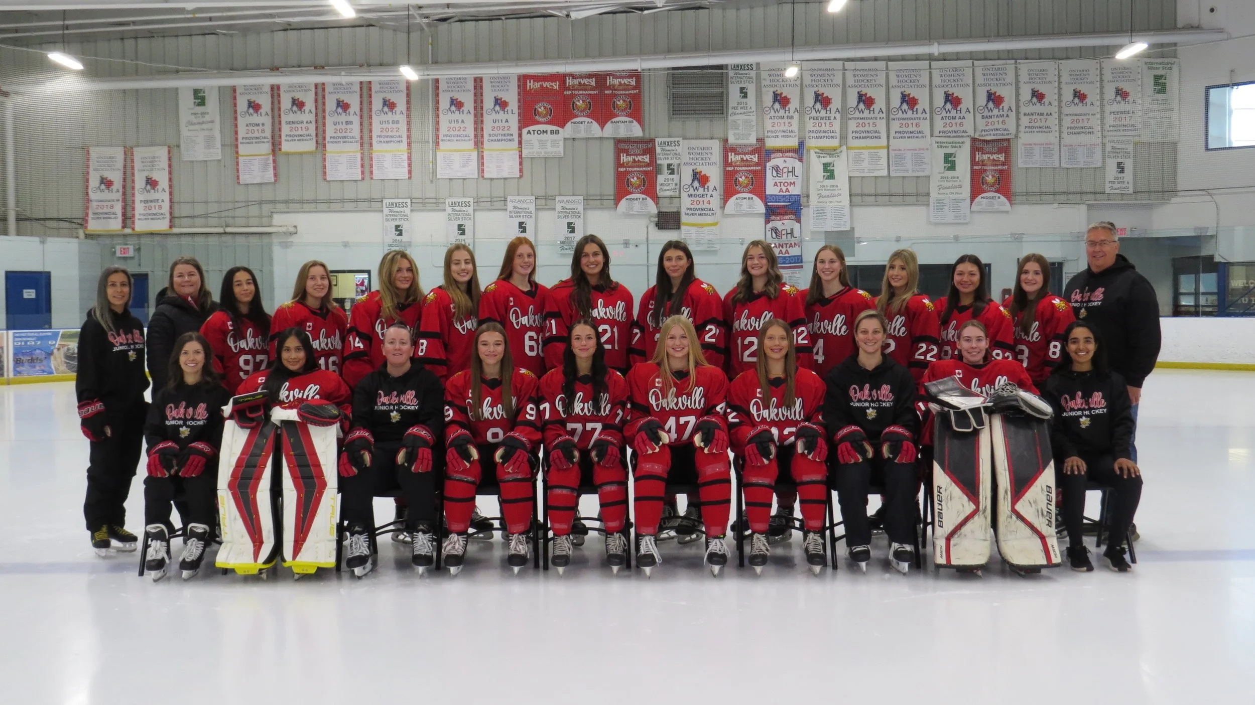Women's hockey team posing on ice rink with red jerseys and banners in the background.