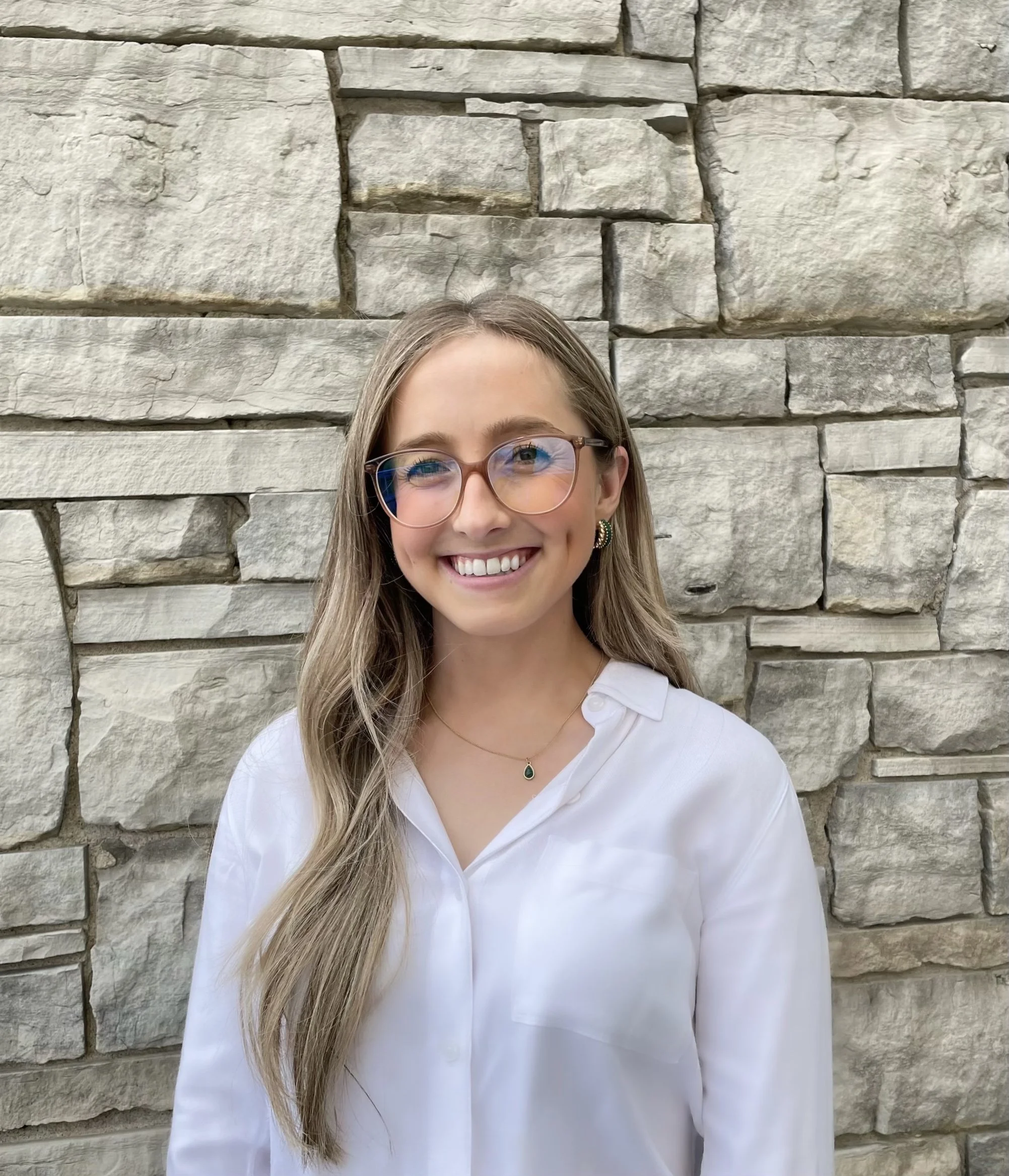 Smiling woman with long hair wearing glasses and a white blouse, standing in front of a stone wall.