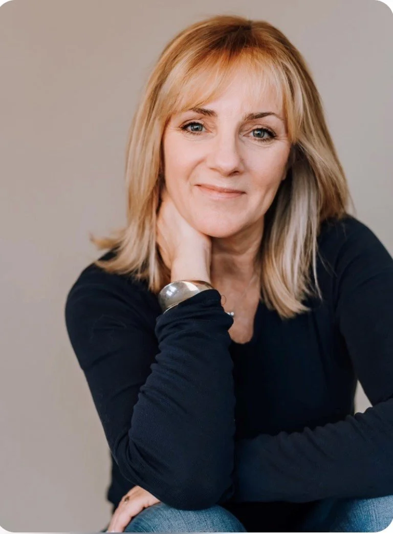 Woman with blonde hair wearing a black long-sleeve top and a silver bracelet, resting her chin on her hand.