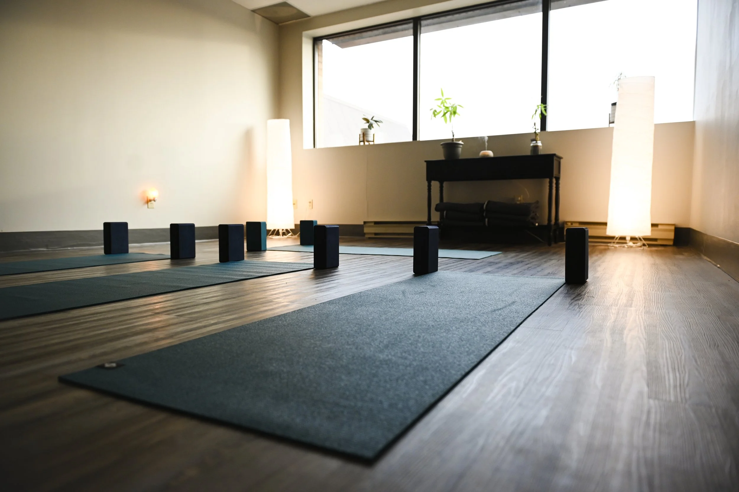 Yoga studio with mats, blocks, and soft lighting, featuring large windows and plants.