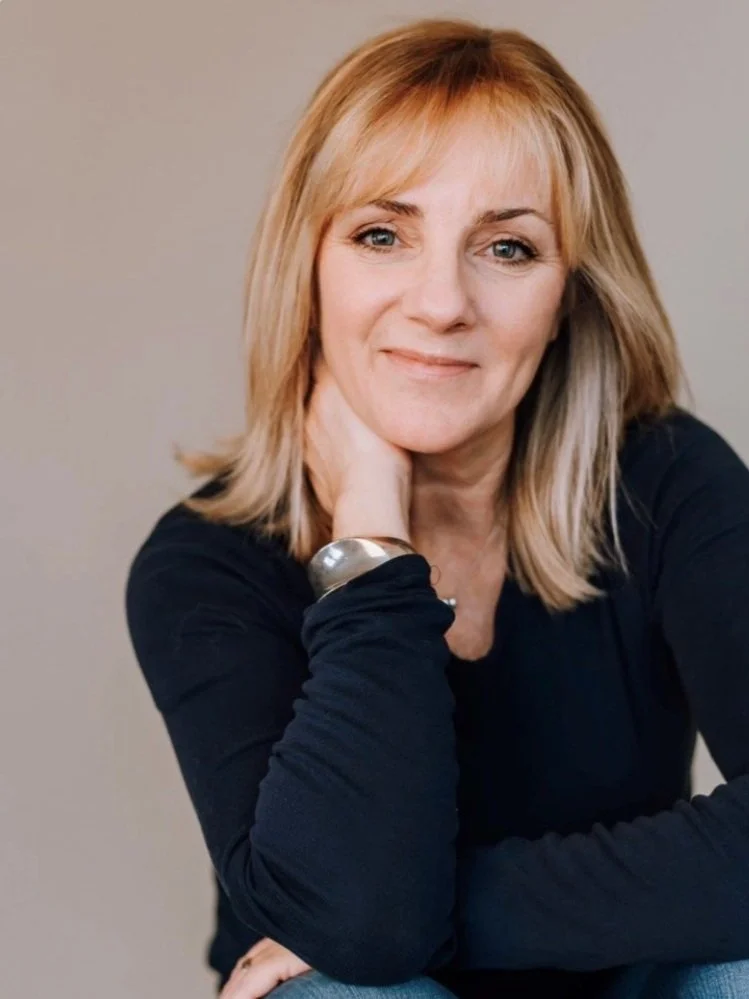 Person with short blonde hair, wearing a black top and silver bracelet, resting chin on hand.
