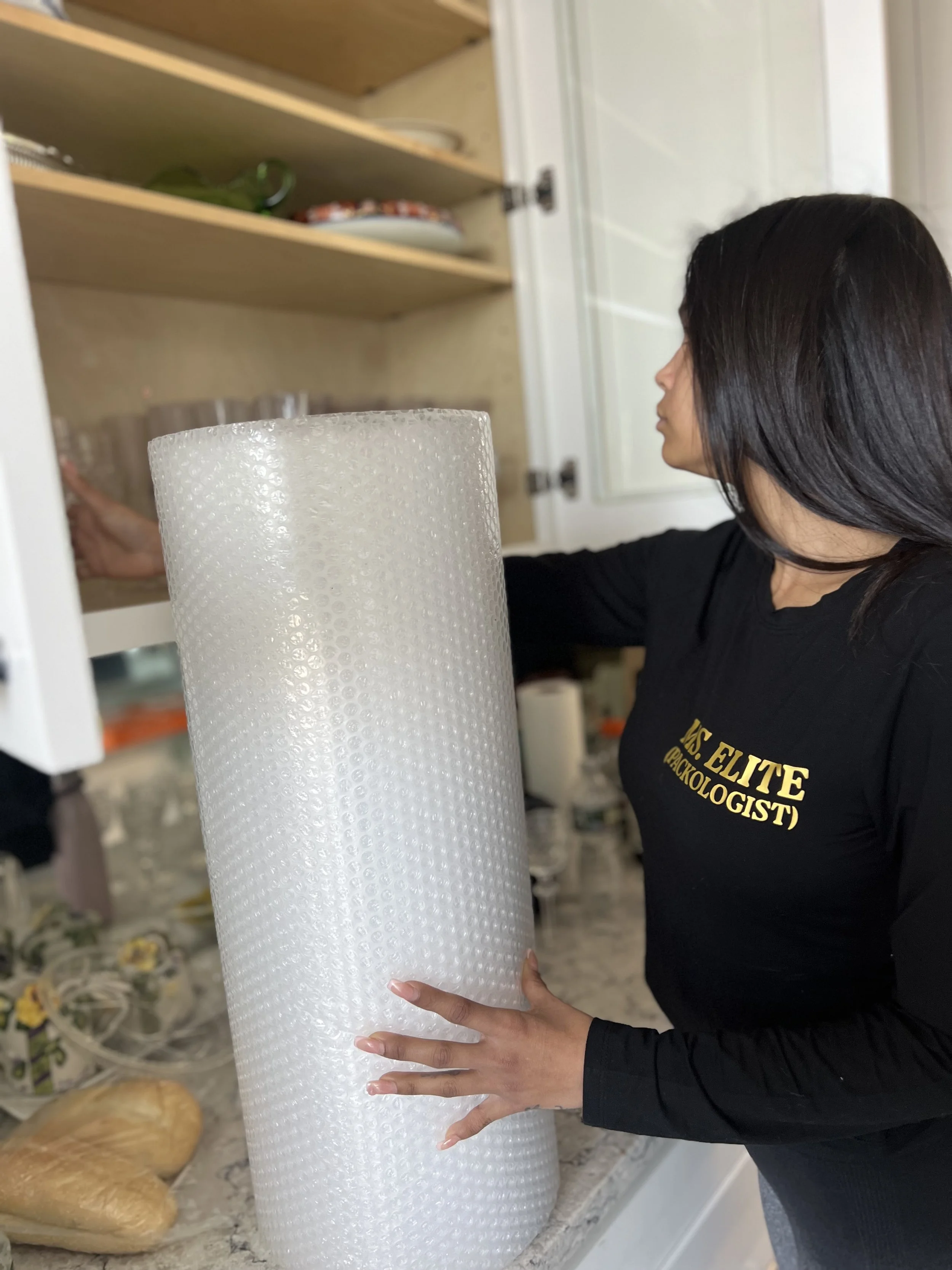A photo of a woman reaching into a cabinet and holding a roll of bubble wrap. She is wearing a shirt that says mr. elite packologist.