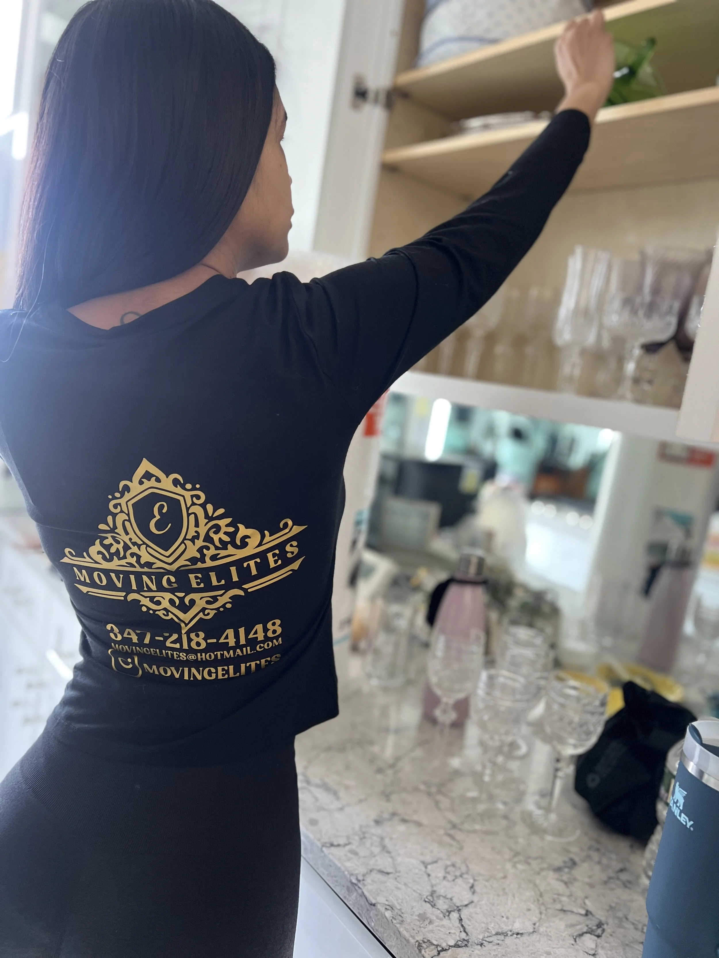 Photo of a woman with black long hair reaching into a kitchen cabinet in order to pack a box with the items. She is wearing a shirt that says moving elites on the back.