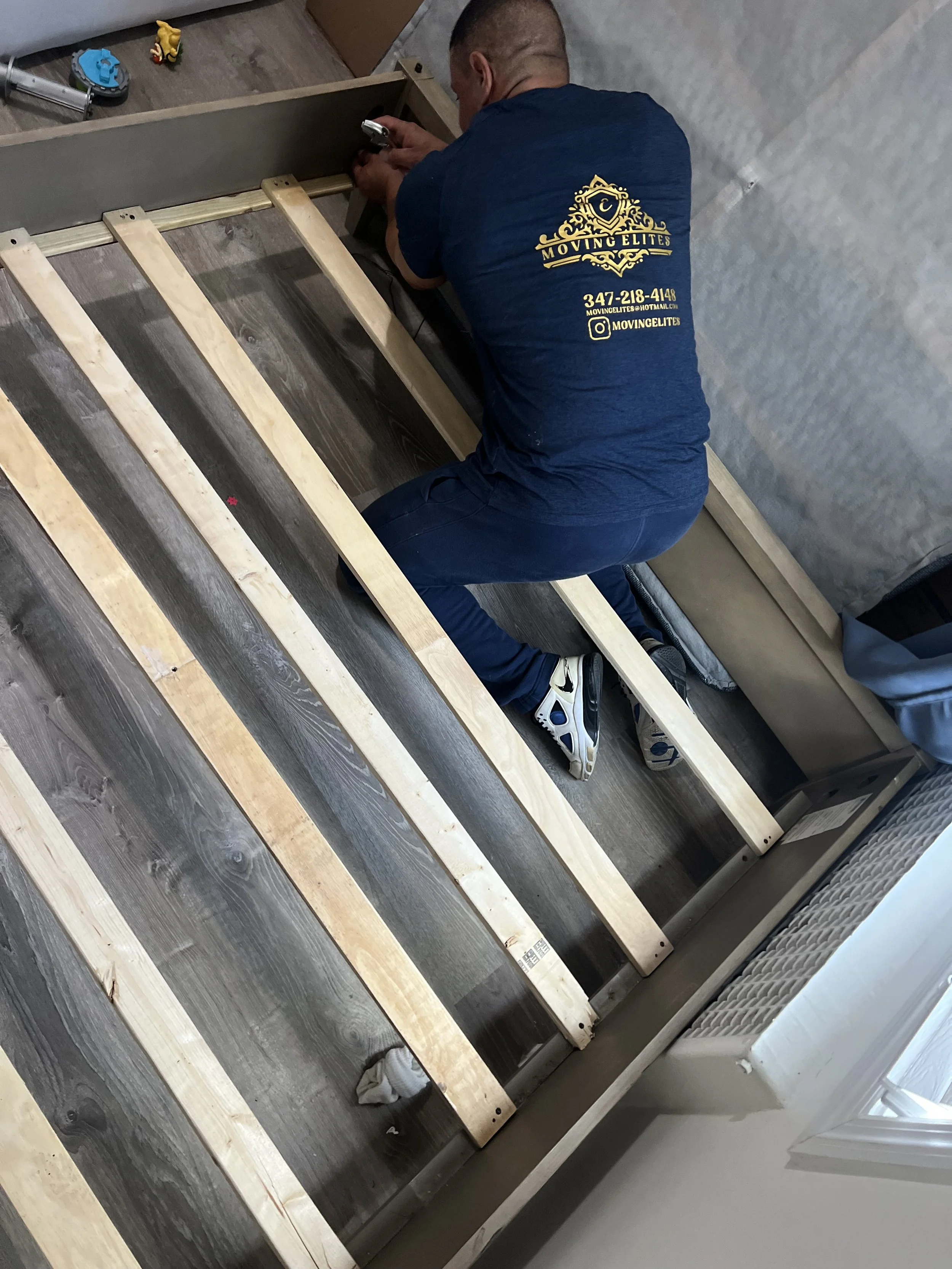 A photo of a man assembling a bed frame. He is wearing a blue shirt that says moving elites.