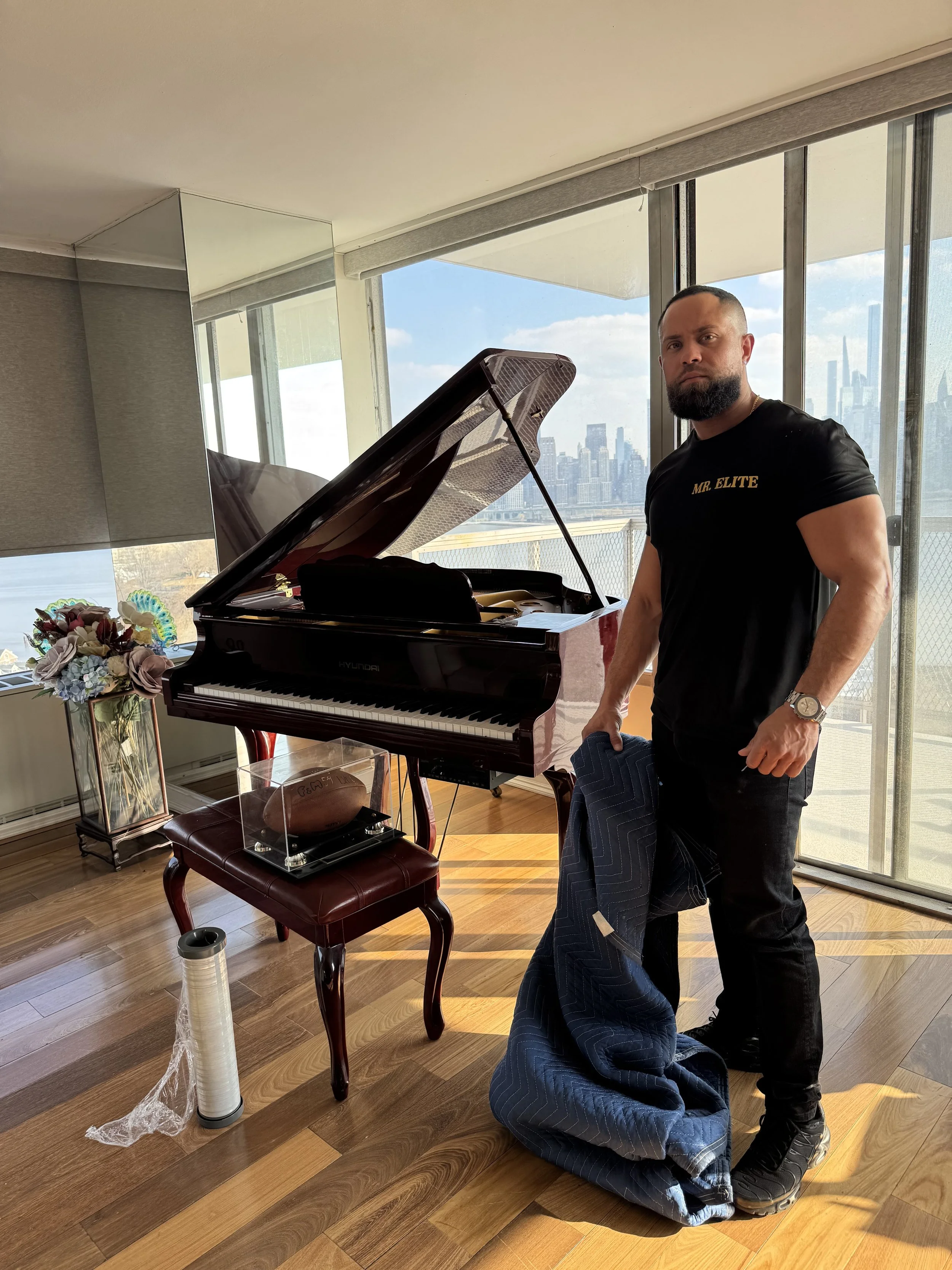 Image of a man holding a moving blanket standing in front of piano. He is wearing a black shirt that say mr. elite
