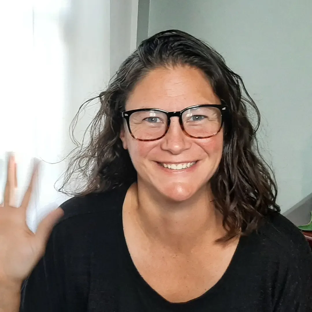 A woman with curly brown hair, wearing glasses and a black shirt, smiling and waving at the camera indoors.