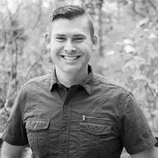 A smiling young man with short hair, wearing a button-up shirt, standing outdoors with a blurred natural background.