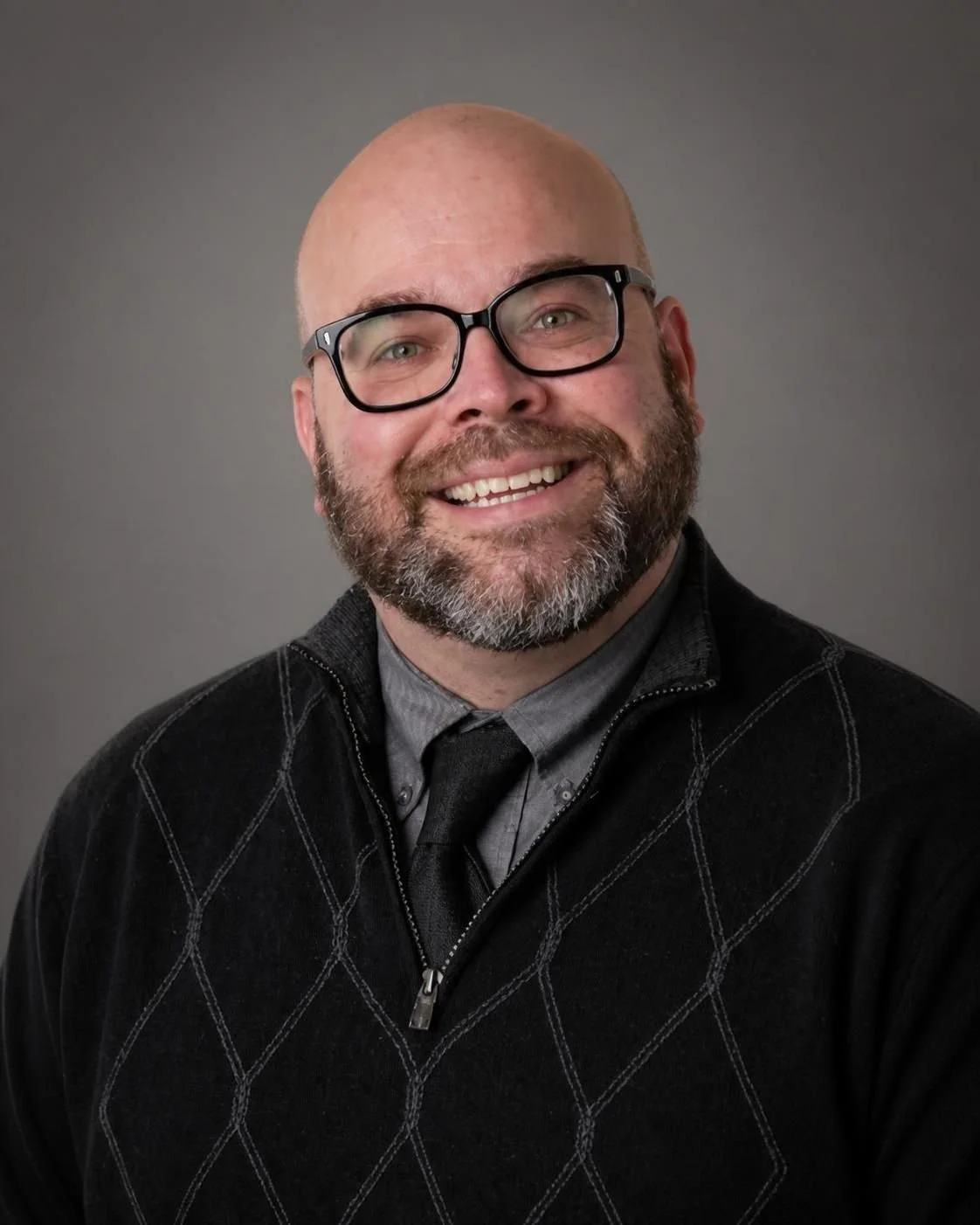 A smiling man with a beard and glasses, wearing a black sweater over a gray shirt and gray tie, against a plain white background.
