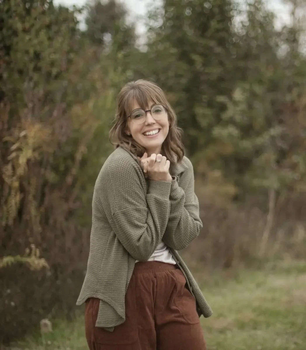 A young woman with glasses and shoulder-length brown hair wearing a green cardigan, white shirt, and brown pants, smiling outdoors near trees.