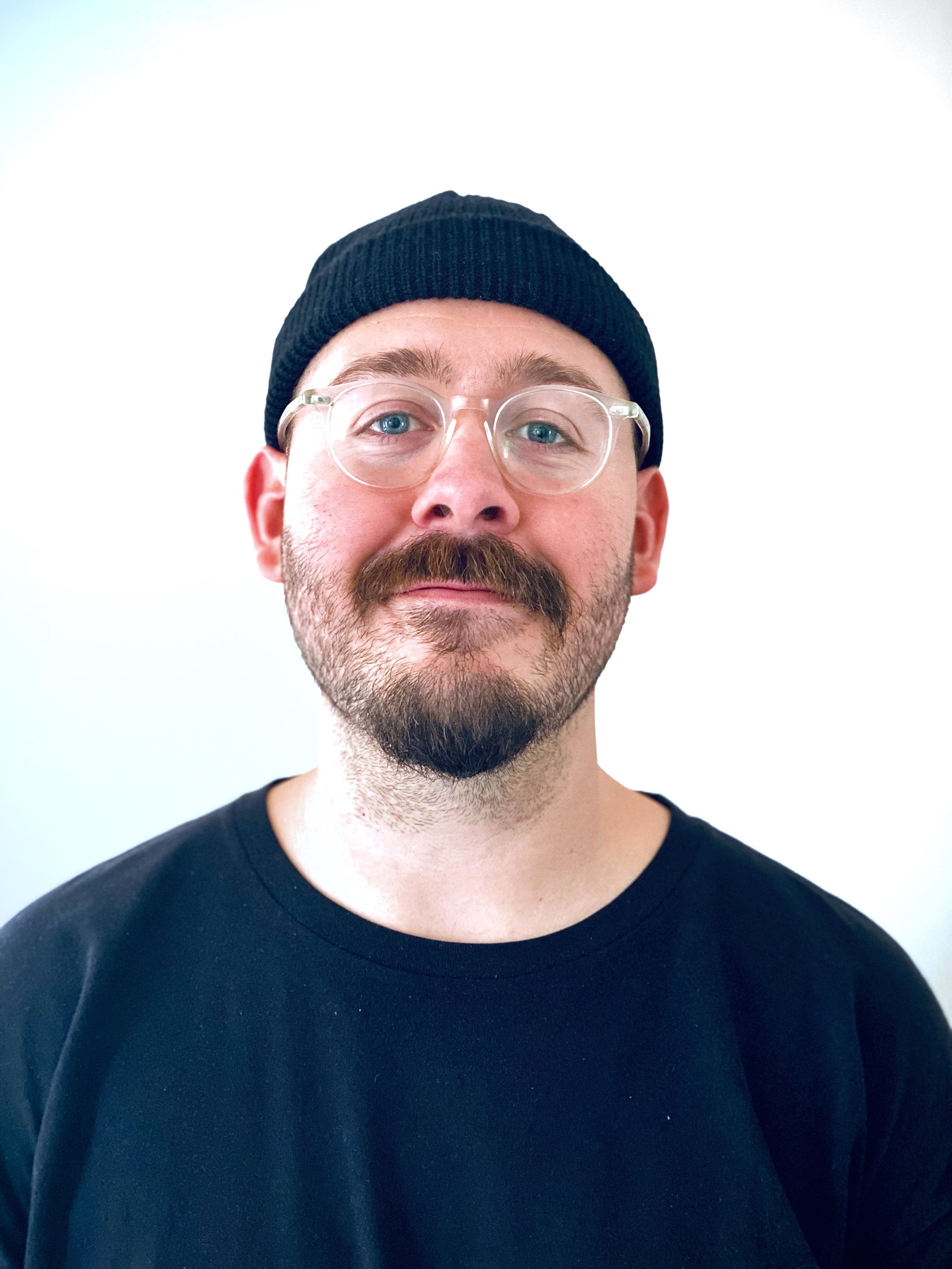 A man with glasses and a beard wearing a black beanie and black shirt, looking directly at the camera against a plain white background.